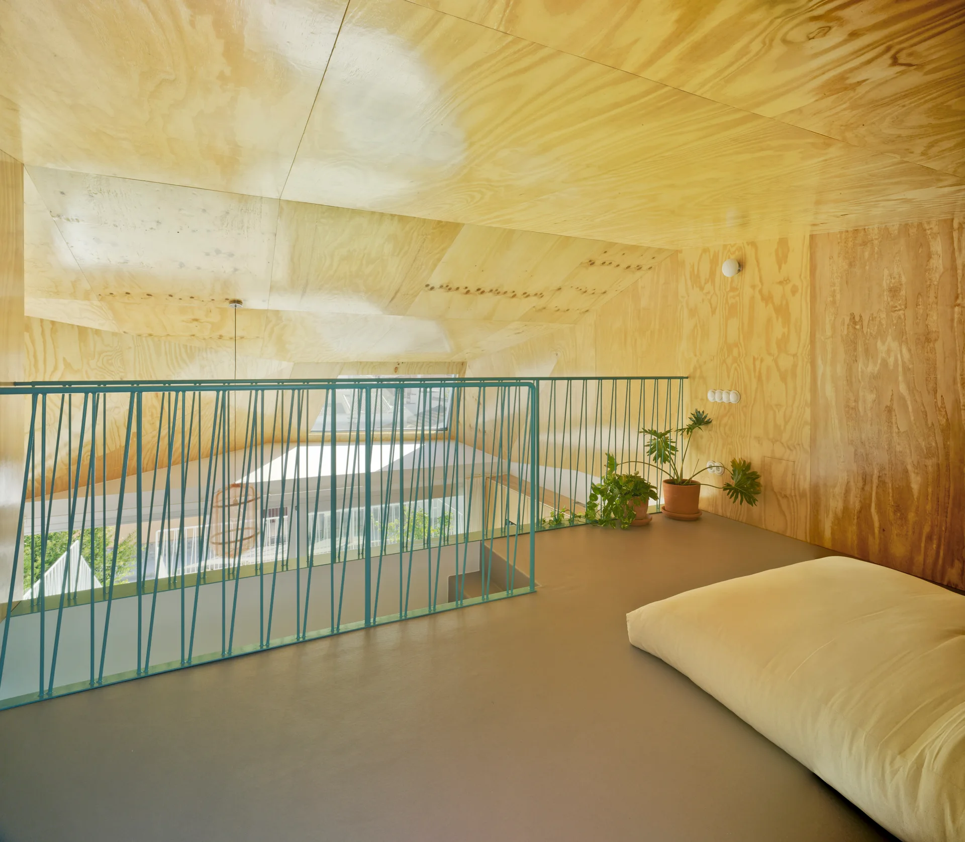 A minimalist loft bedroom features plywood-paneled walls and ceiling, a flat gray floor, a simple floor bed, and a distinctive teal metal safety railing.