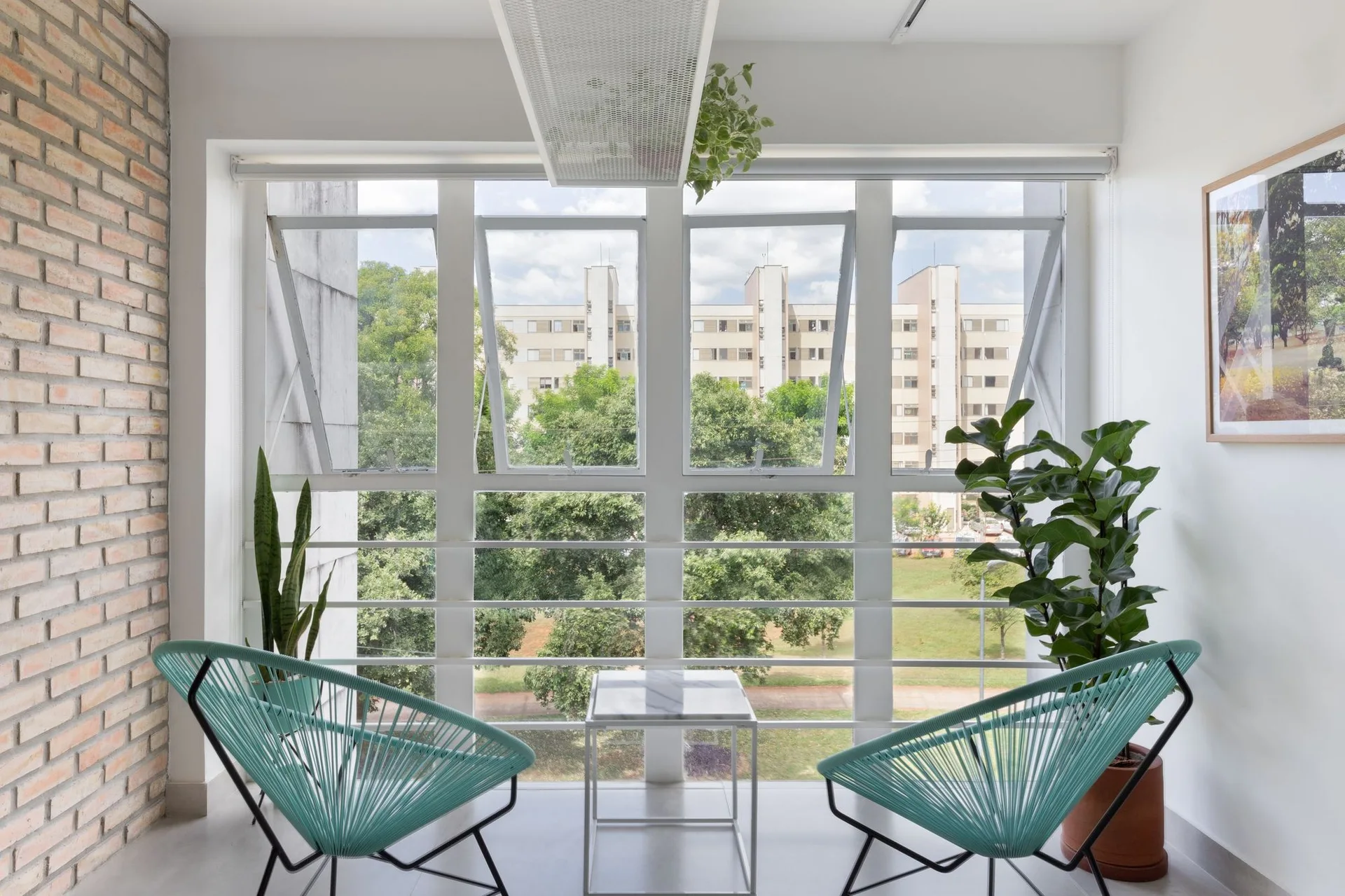 Two turquoise woven string chairs face a small marble-topped table in front of a large window, next to an exposed brick wall.