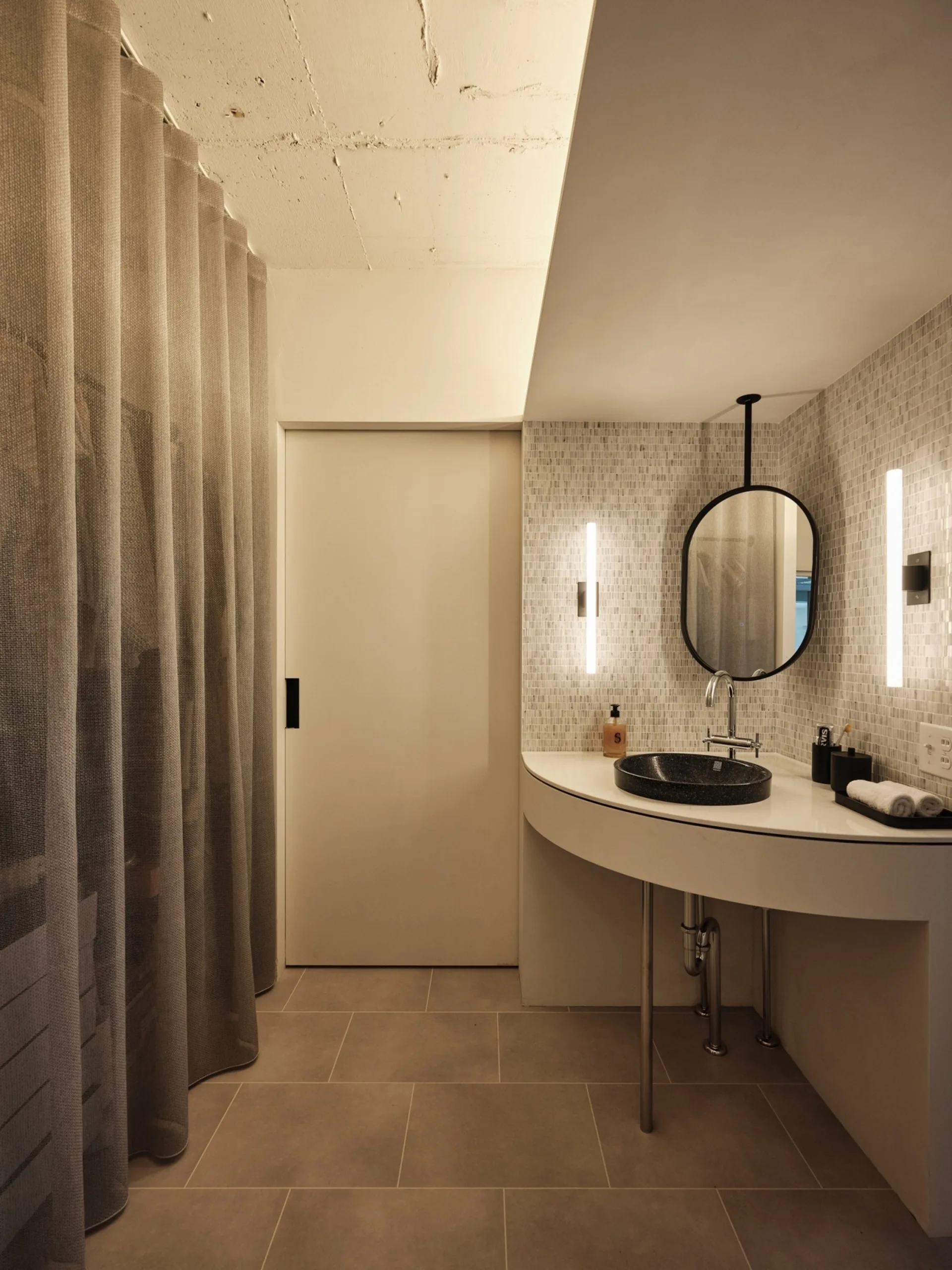 A modern bathroom features a white curved vanity with a dark round vessel sink, an oval black-framed mirror, and vertical sconces against a tiled wall.