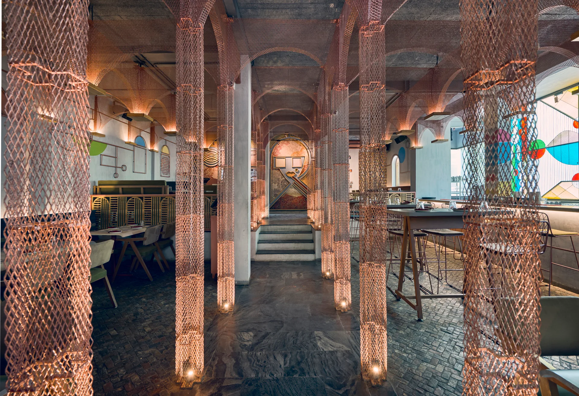 A restaurant dining room featuring copper metal mesh columns that form arches under a grey ceiling, lit by warm base lights on a stone floor.