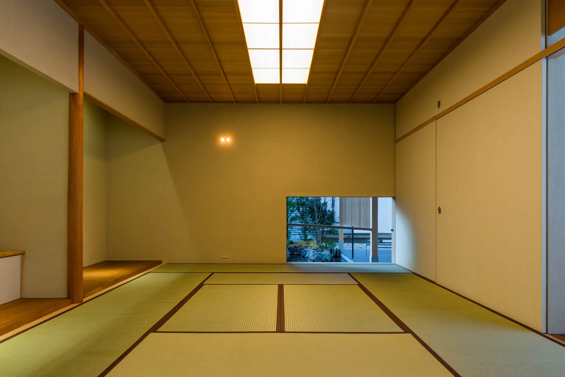 A minimalist Japanese room features green tatami floor mats, a wood grid ceiling, and a low rectangular window looking out to a garden.