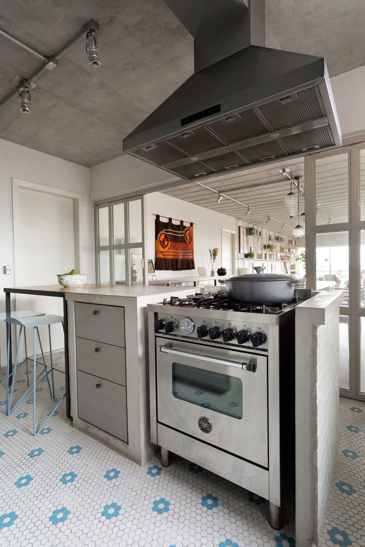 An industrial kitchen features a stainless steel range under a metal hood, flanked by concrete counters, set on a blue-and-white hexagonal mosaic tile floor.