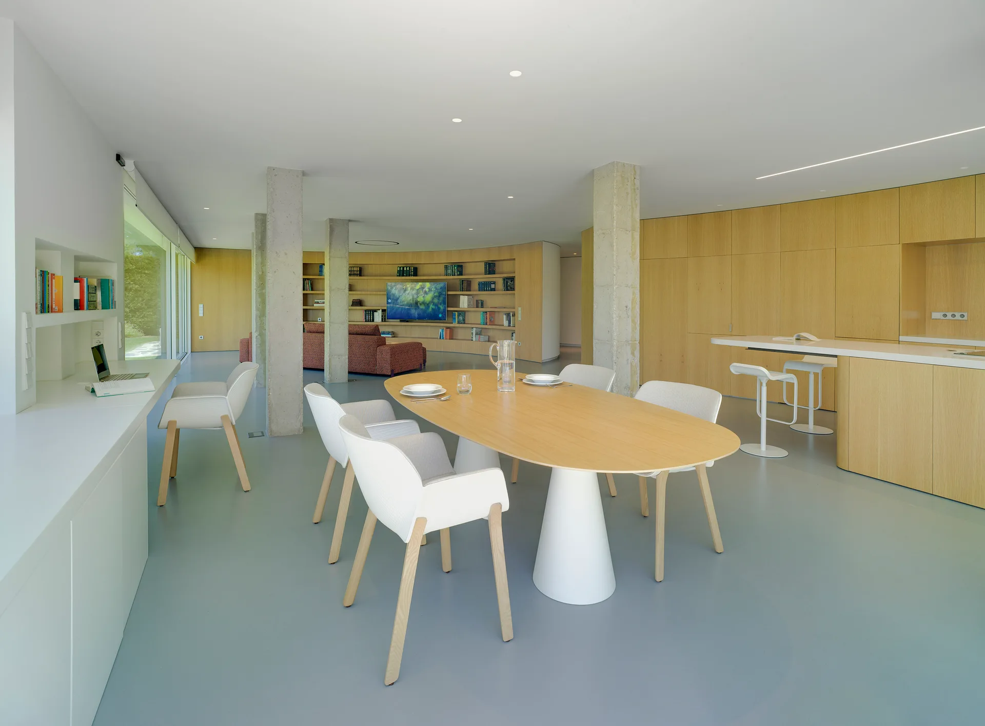An open-concept dining room features an oval wood table, white chairs, exposed concrete columns, and extensive light wood wall paneling.