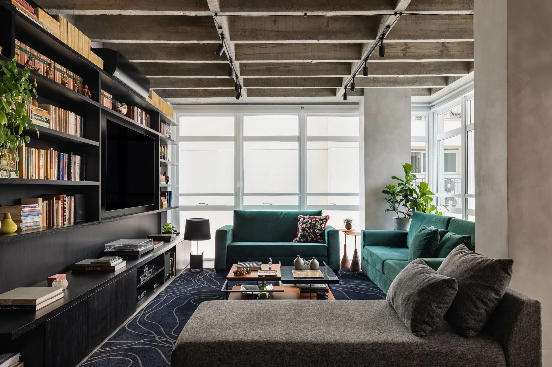 A modern apartment living room features emerald green velvet sofas, a black library wall with a television, and an industrial exposed concrete ceiling.