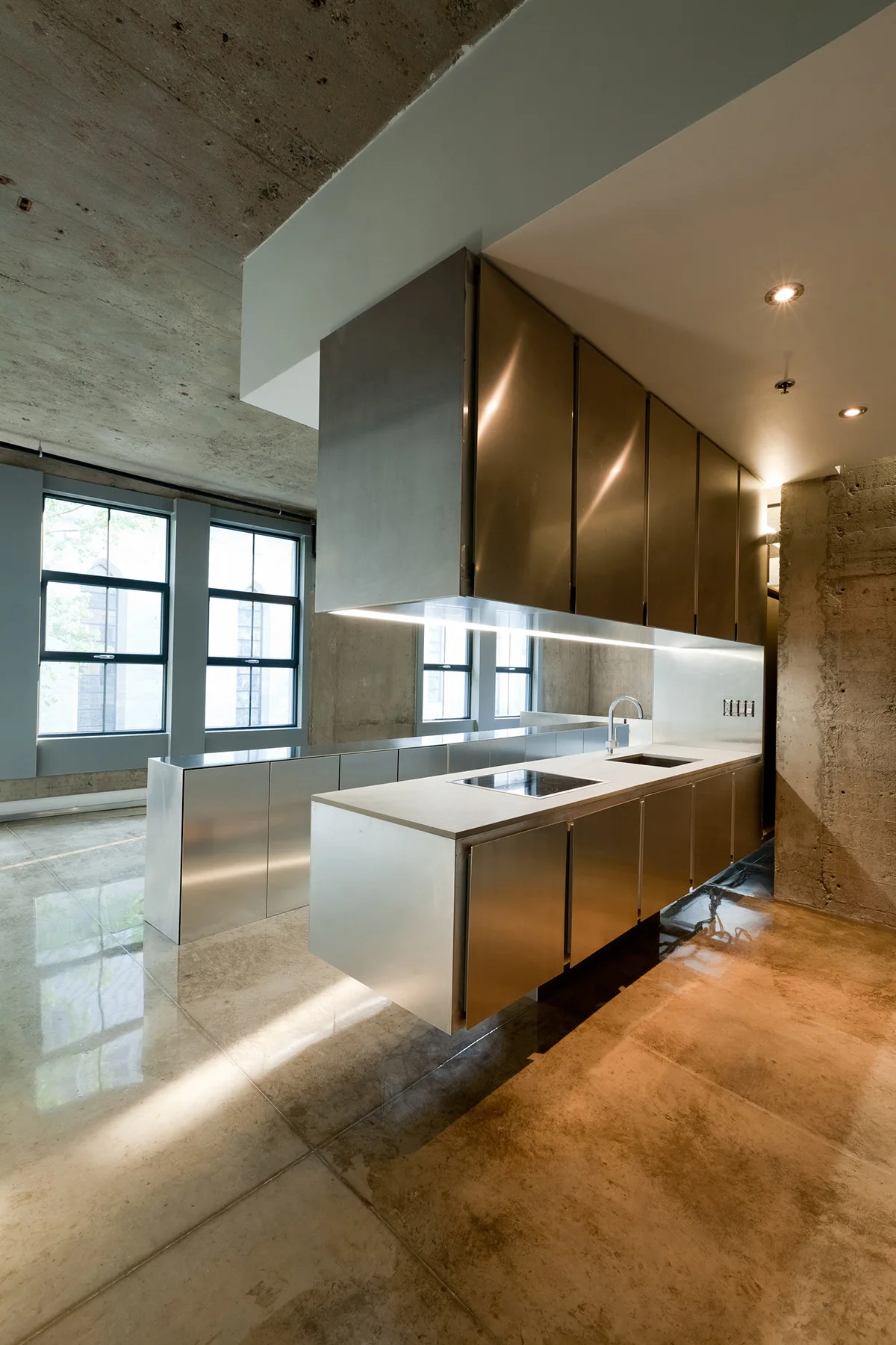 An industrial kitchen features brushed stainless steel cabinets, a floating island, and raw concrete walls and floors.