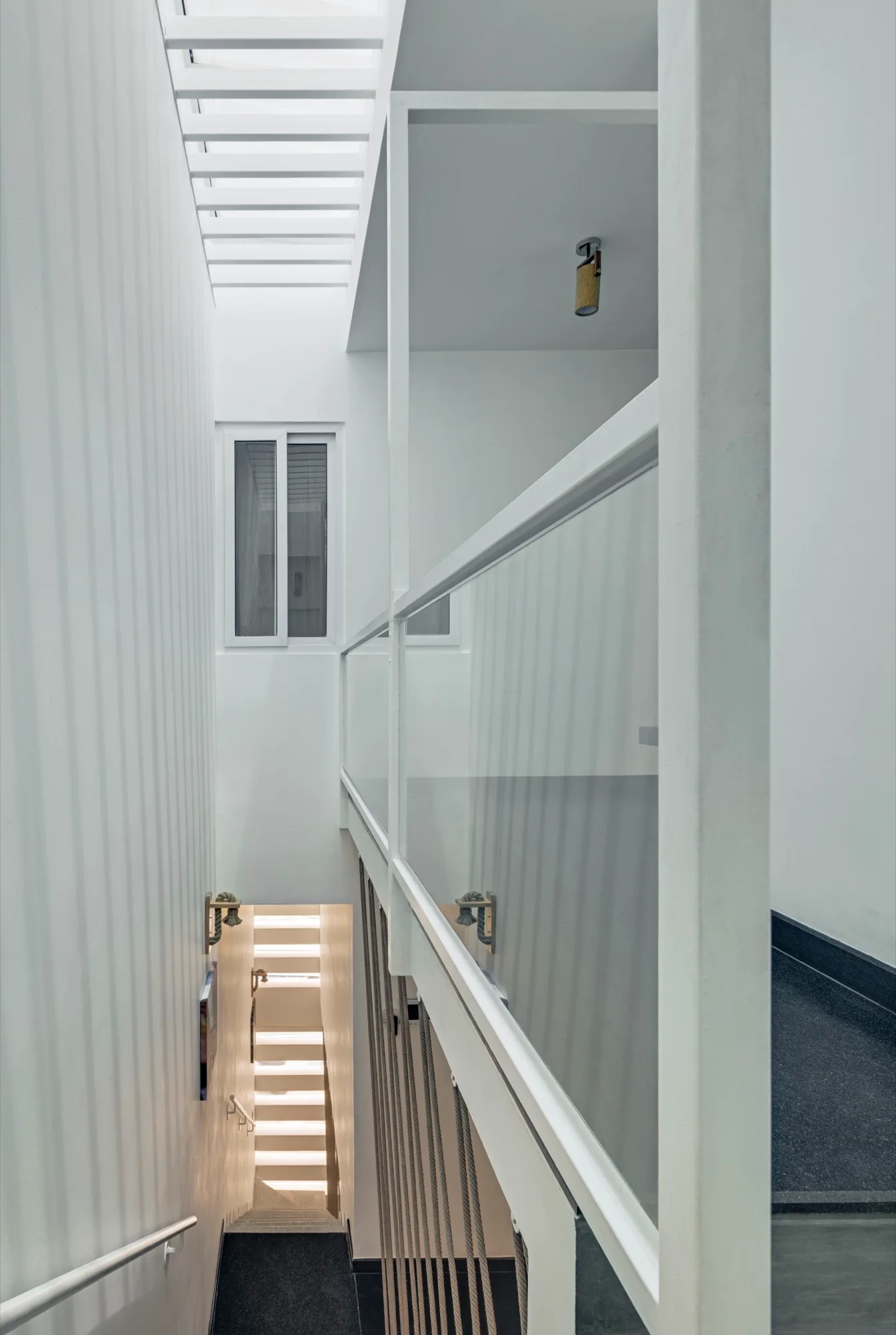 A minimalist multi-level white stairwell features dark grey stairs, a glass railing, and a slatted skylight that casts shadows on the wall.