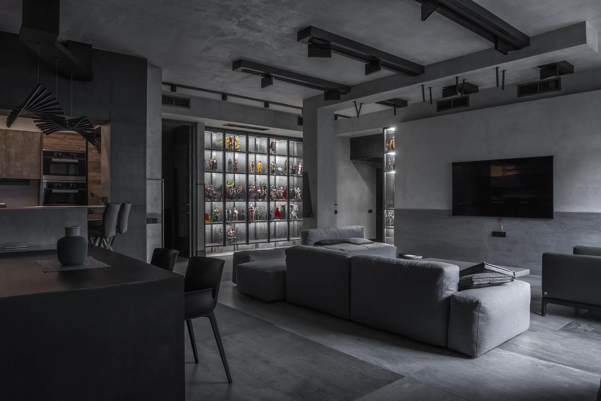 A dark grey living room features a concrete-finish ceiling with track lighting, a matching sectional, and a large backlit display cabinet for figurines.