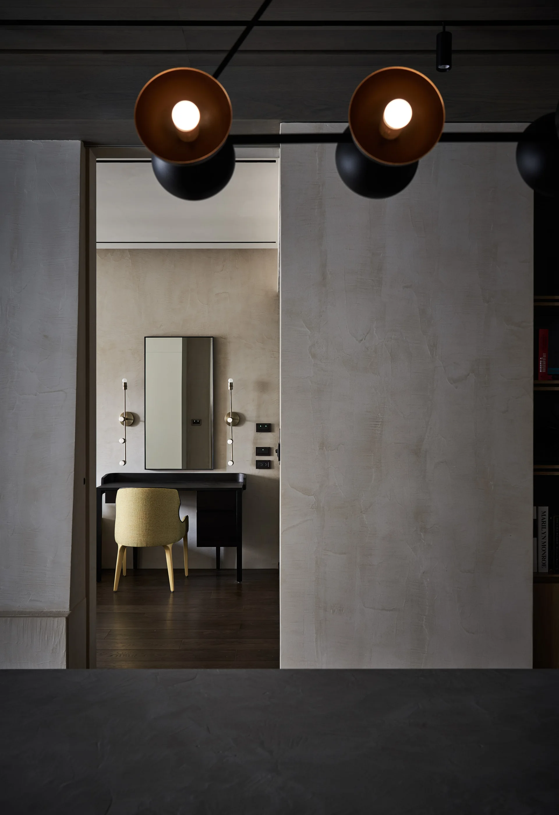 A view through a doorway into a minimalist dressing nook with a dark vanity desk, a yellow chair, and a rectangular mirror flanked by slim wall sconces.