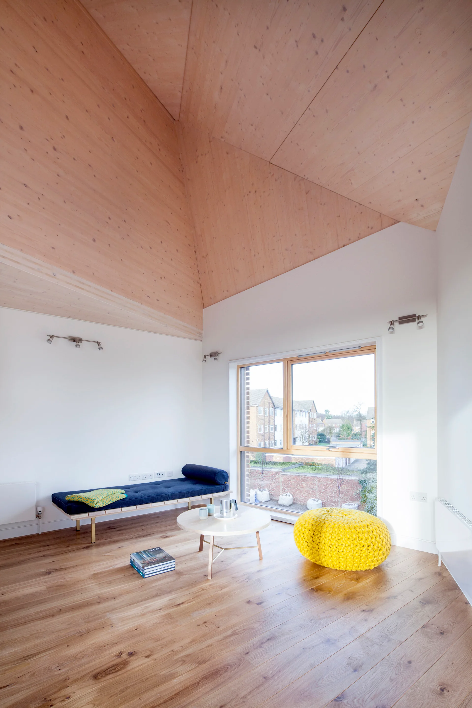 A minimalist living room with a vaulted plywood ceiling, white walls, a navy daybed, a round coffee table, and a bright yellow knit ottoman.