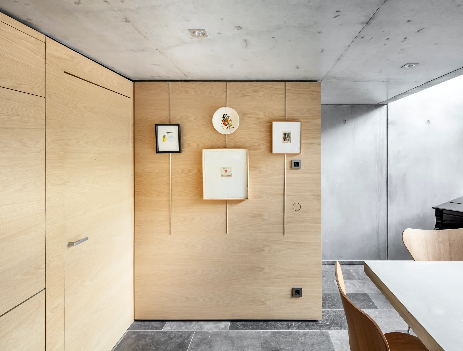 A minimalist dining area features light wood wall paneling with hanging artwork, an exposed concrete ceiling, and grey stone floor tiles.