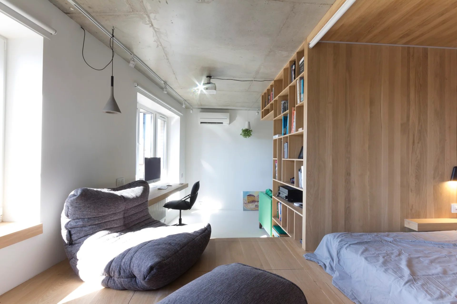 A modern studio bedroom featuring a split-level wood floor, a built-in wooden bookshelf divider, grey lounge chairs, and an exposed concrete ceiling.