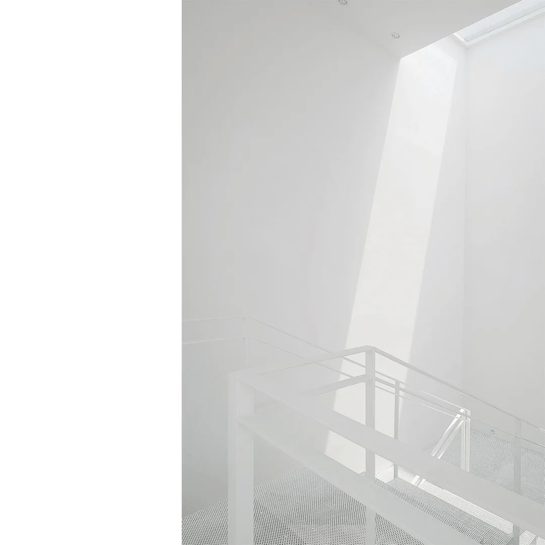 A minimalist all-white metal staircase with mesh treads is illuminated by a beam of natural sunlight from an overhead skylight.
