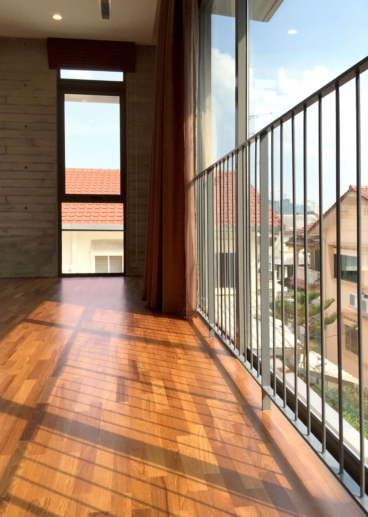An indoor space features warm wood plank flooring, a textured concrete wall, and a large window looking out onto a metal balcony railing.