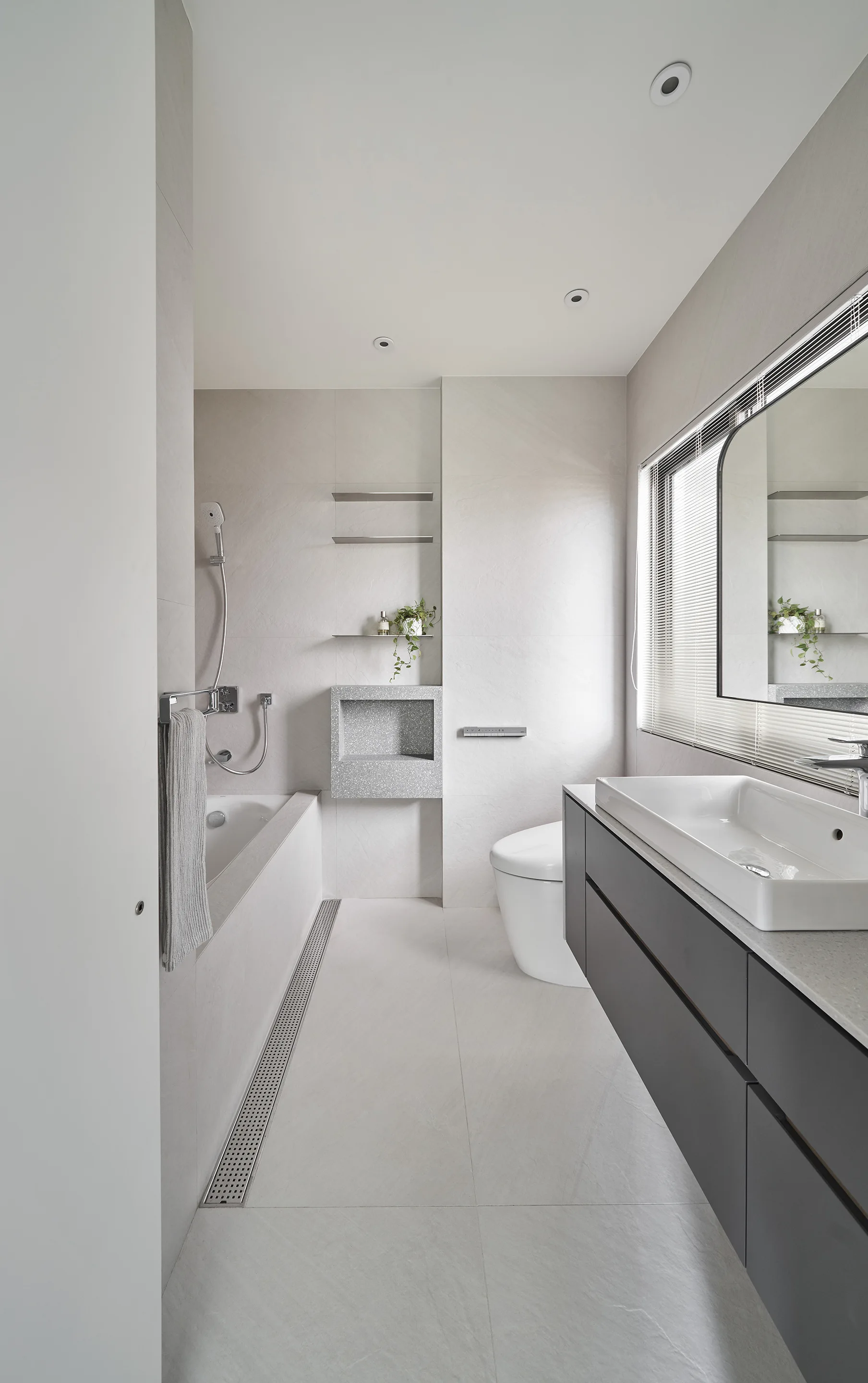 A minimalist grey bathroom featuring a dark grey floating vanity, a built-in white bathtub, and a long linear drain in the tiled floor.