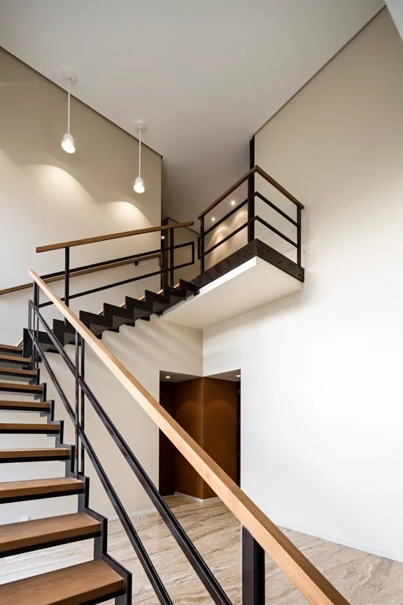 A modern floating staircase with wood treads, black metal geometric railings, and a wood handrail rises toward a beige hallway landing with pendant lights.