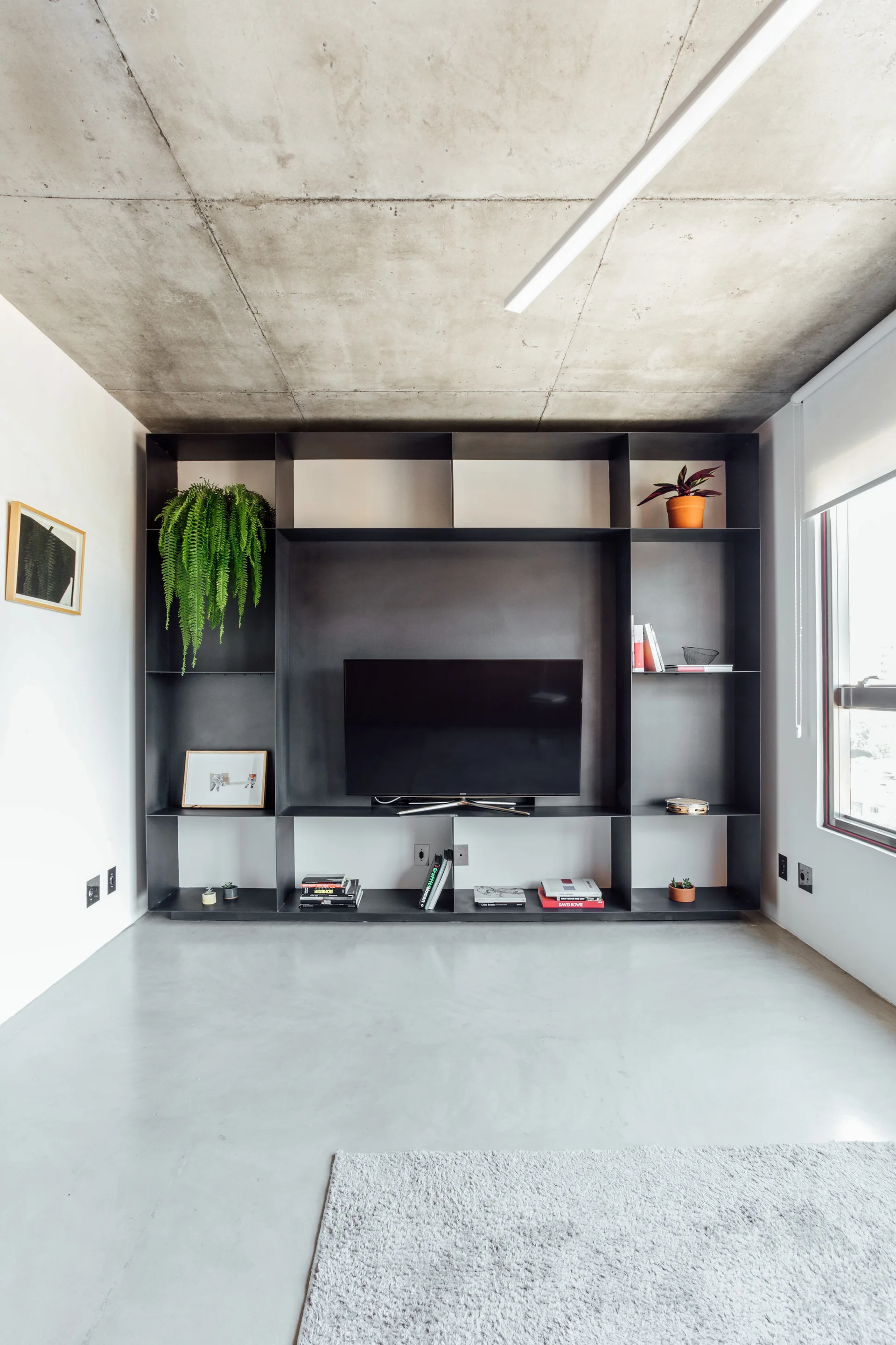 A minimalist living room features a custom black metal shelving unit with a TV, set between a raw concrete ceiling and polished concrete floor.