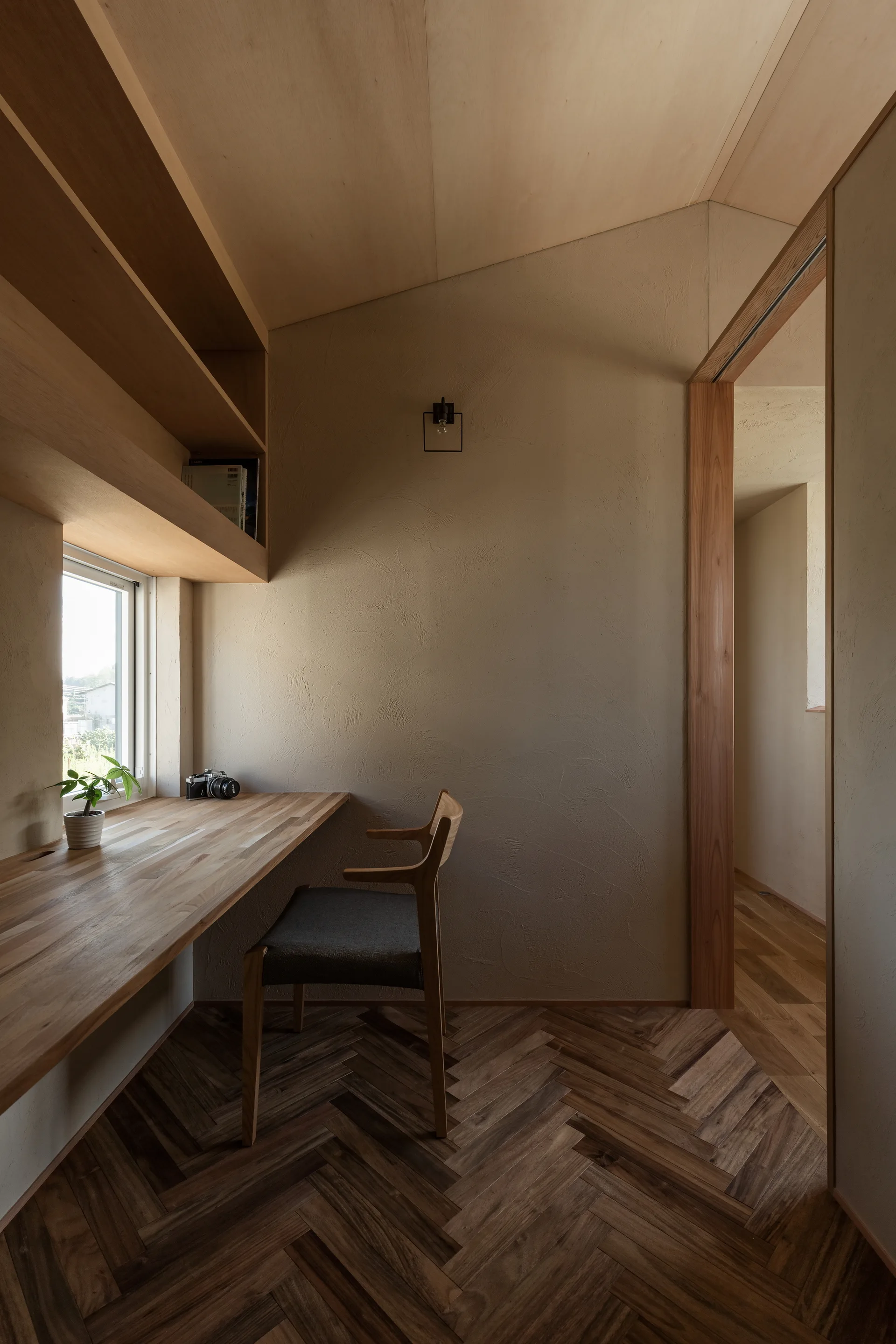 A minimalist home office featuring a built-in wooden desk, a wooden chair, textured plaster walls, and a warm herringbone wood floor.