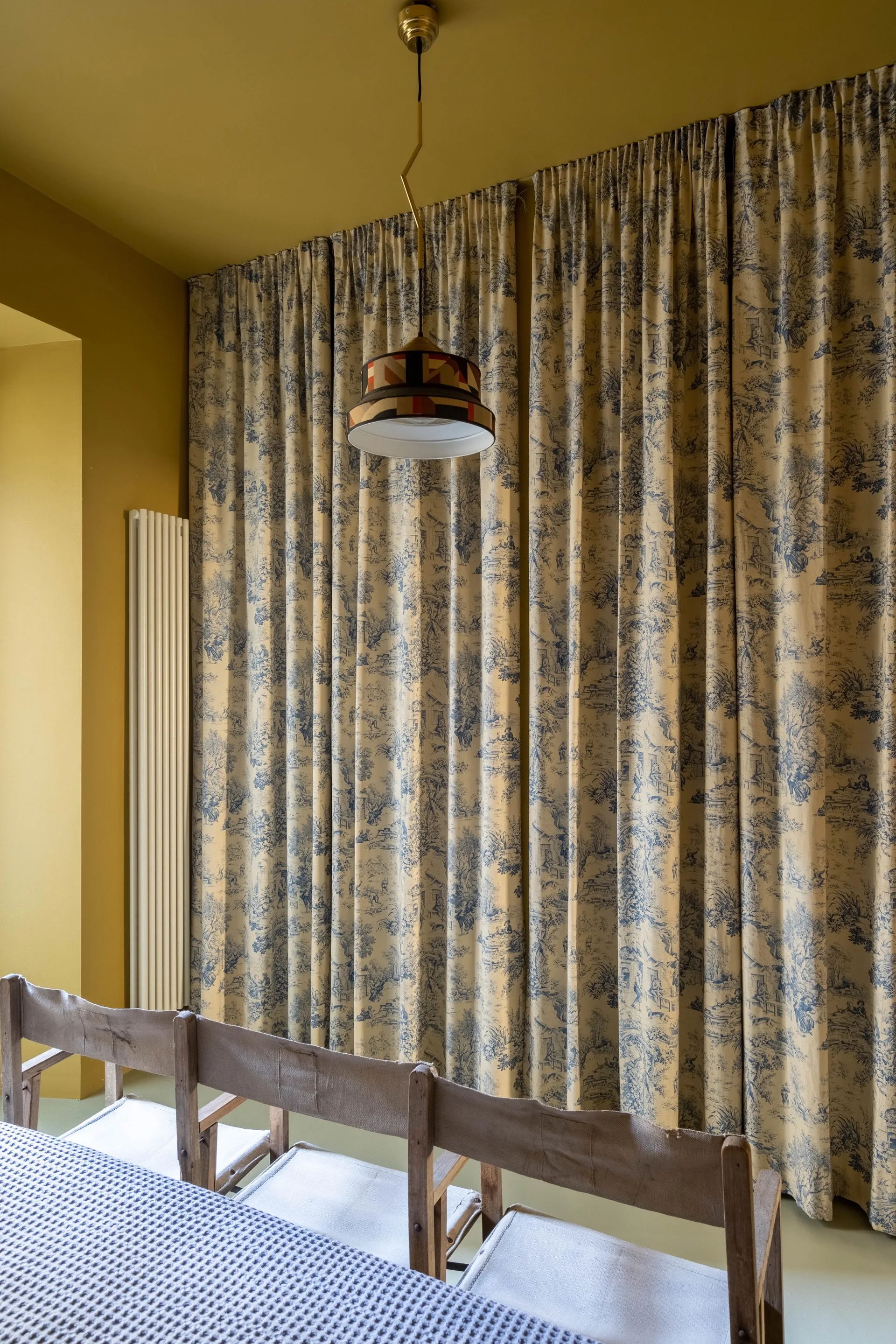 Floor-to-ceiling blue and white toile curtains hang in an ochre-painted dining room with a geometric pendant lamp and simple wooden chairs.
