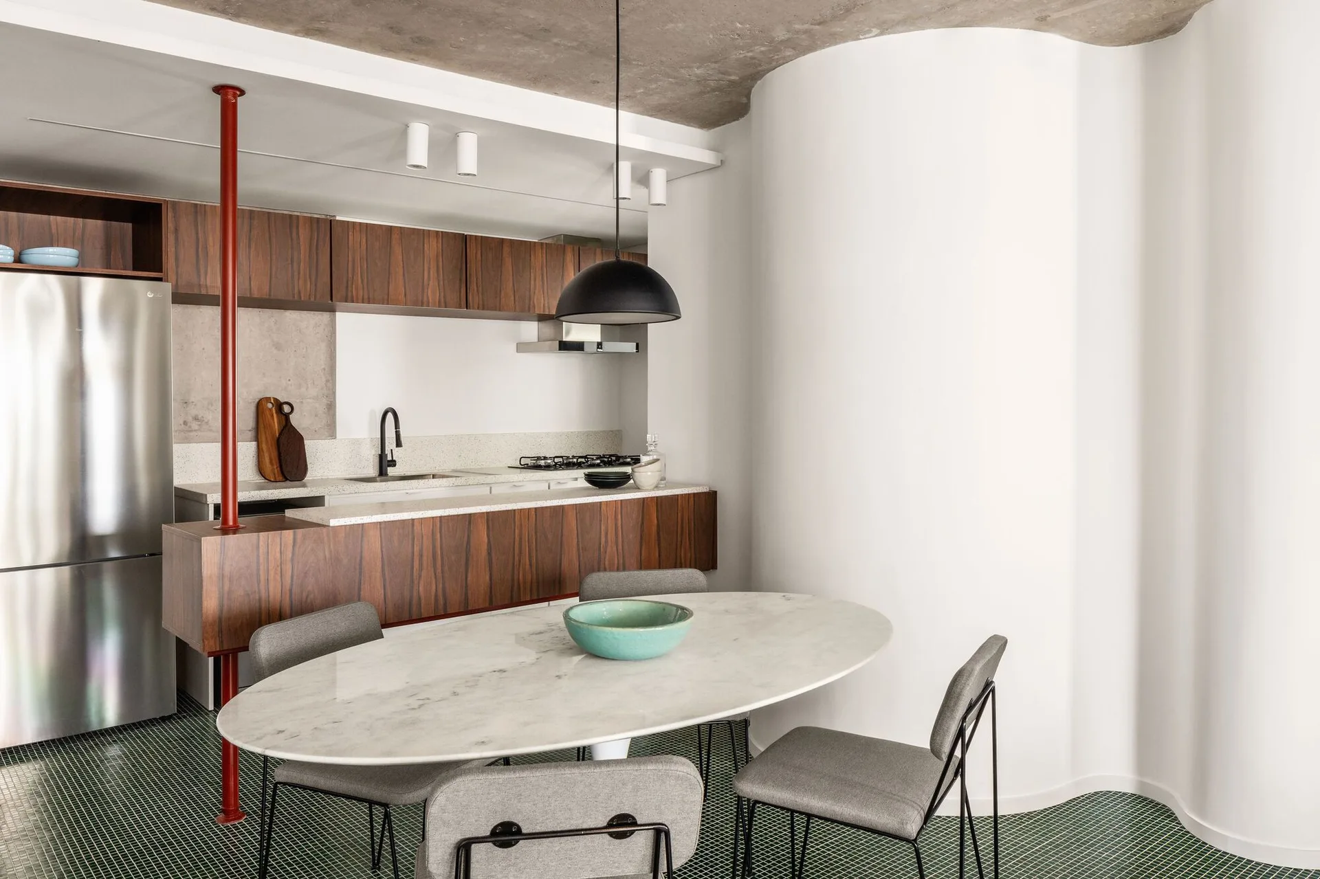 A contemporary kitchen and dining space with an oval marble table, dark wood cabinets, a curved white wall, and green mosaic tile flooring.