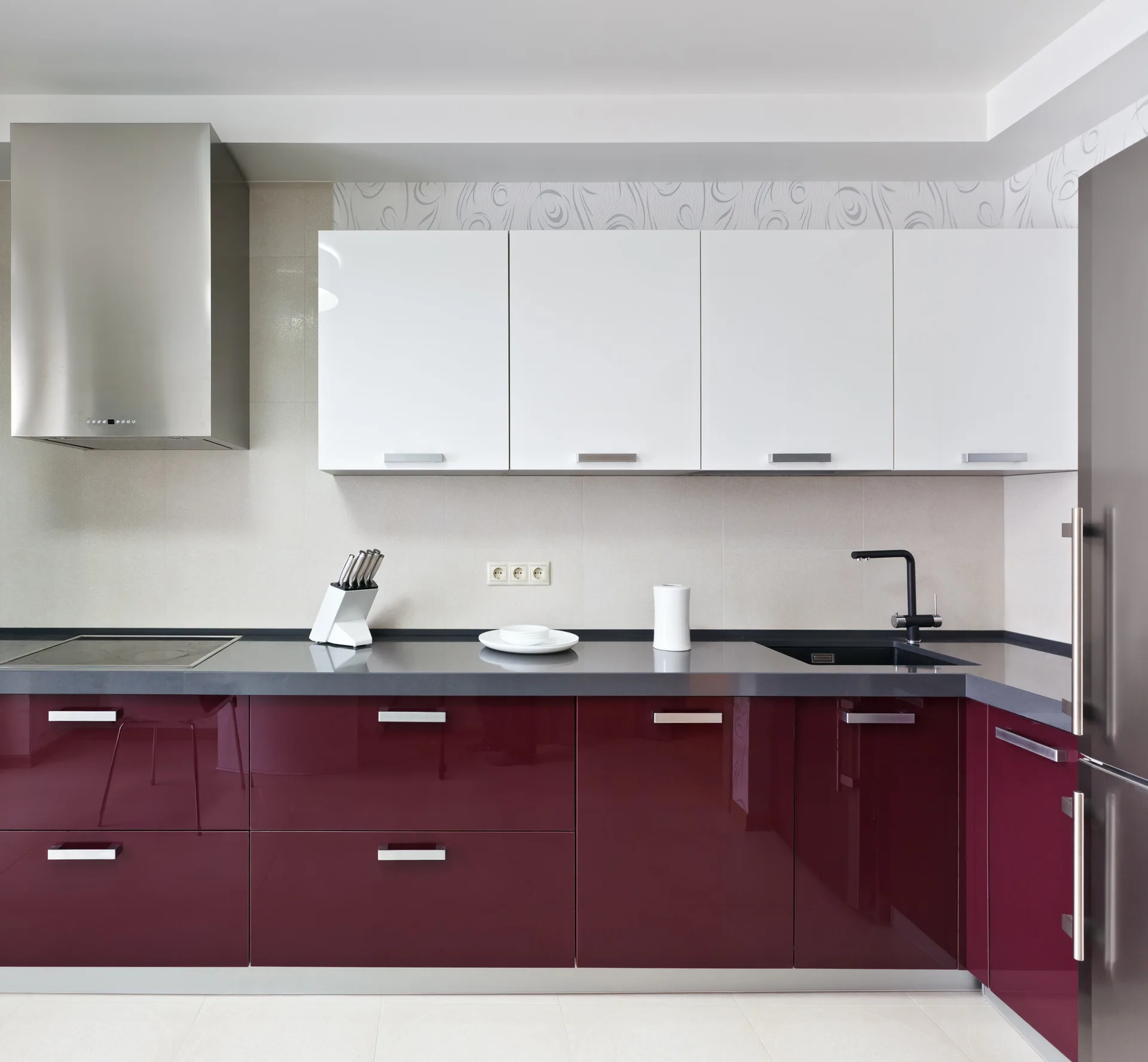 A modern kitchen features glossy maroon lower cabinets, white upper cabinets, a dark grey countertop, and a stainless steel range hood.