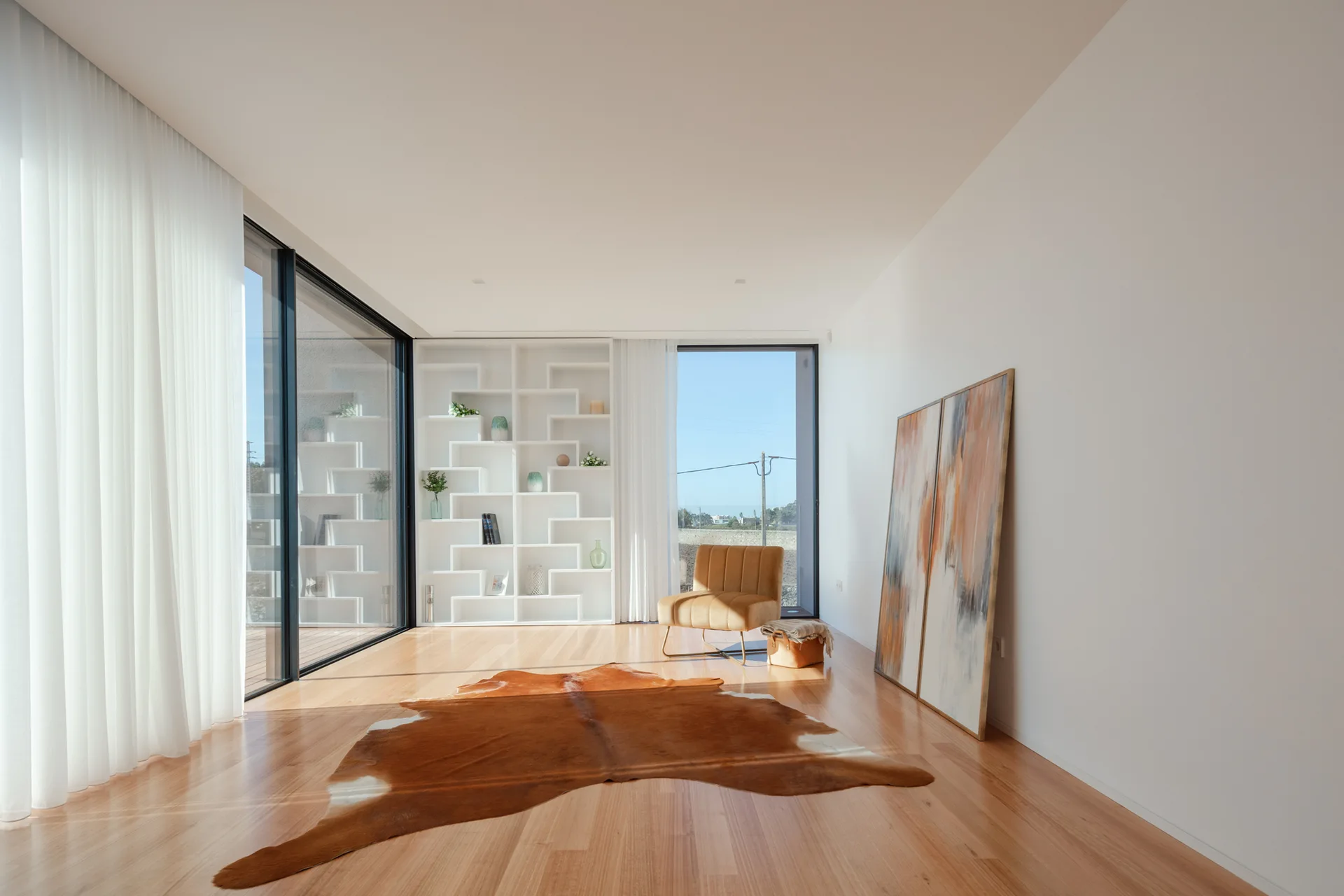 A minimalist white living room with light wood floors, an asymmetrical built-in bookshelf, a cowhide rug, and two paintings leaning against the wall.