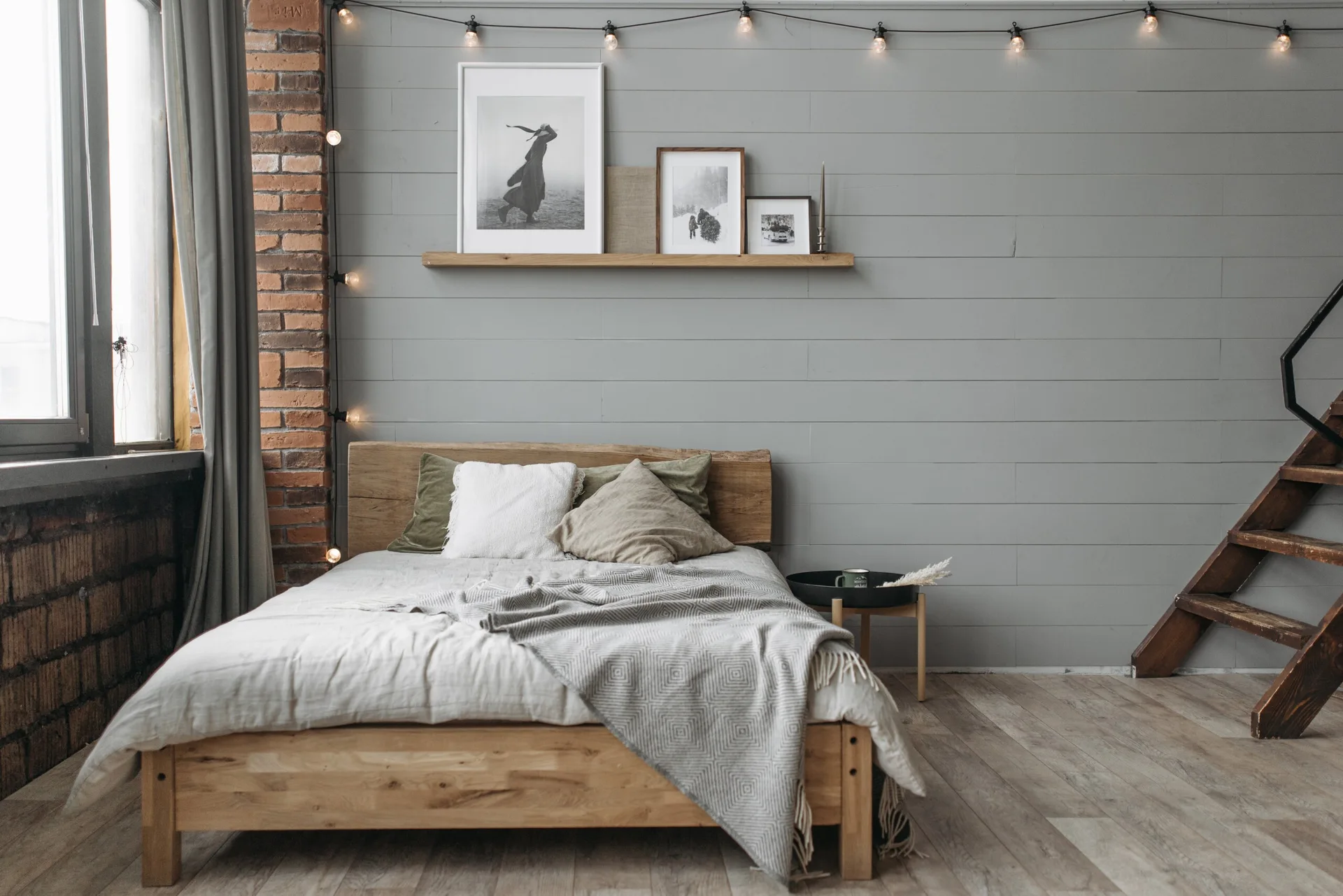 A rustic bedroom features a wooden bed frame, a grey wood plank wall with string lights, and a small brick pillar on the left.