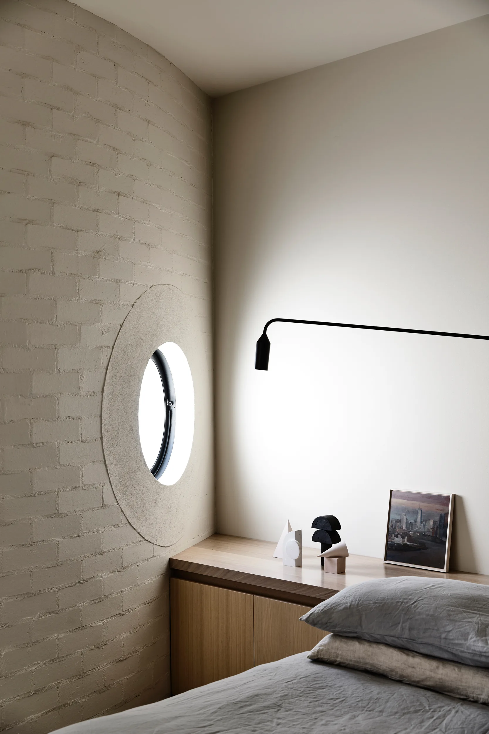 A minimalist bedroom corner featuring a circular window set in a painted beige brick wall, next to a wooden built-in ledge and grey bed linens.