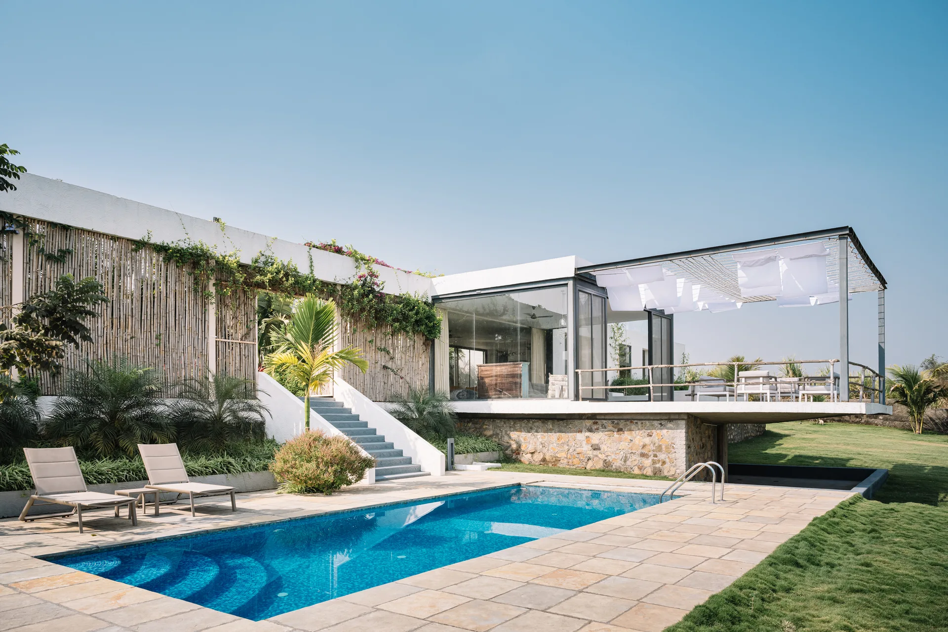 A rectangular blue swimming pool sits next to a modern white house with a stone foundation, a bamboo screen facade, and an outdoor staircase.