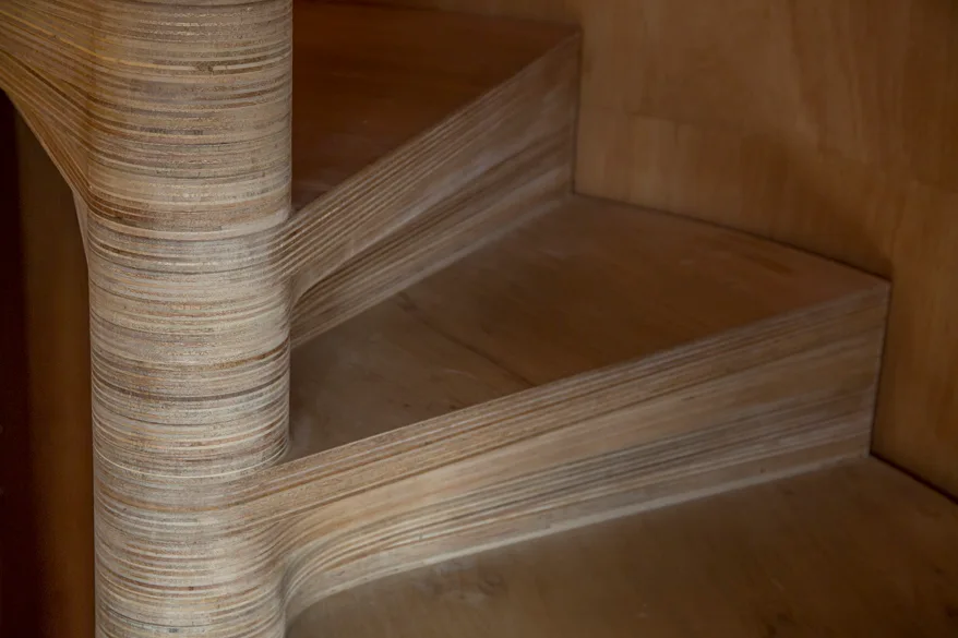 A close-up shot of a curved plywood staircase showing the detailed laminated wood layers on the steps and central column.