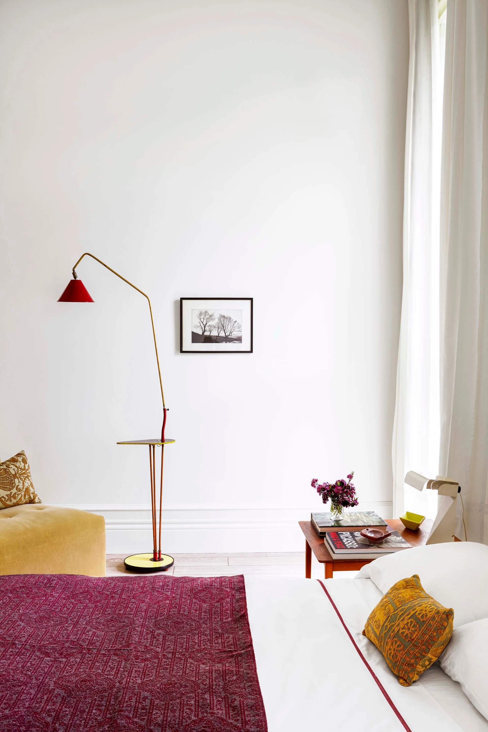 A bright white bedroom features a bed with burgundy bedding, a wooden bedside table, and a slender red and yellow floor lamp.
