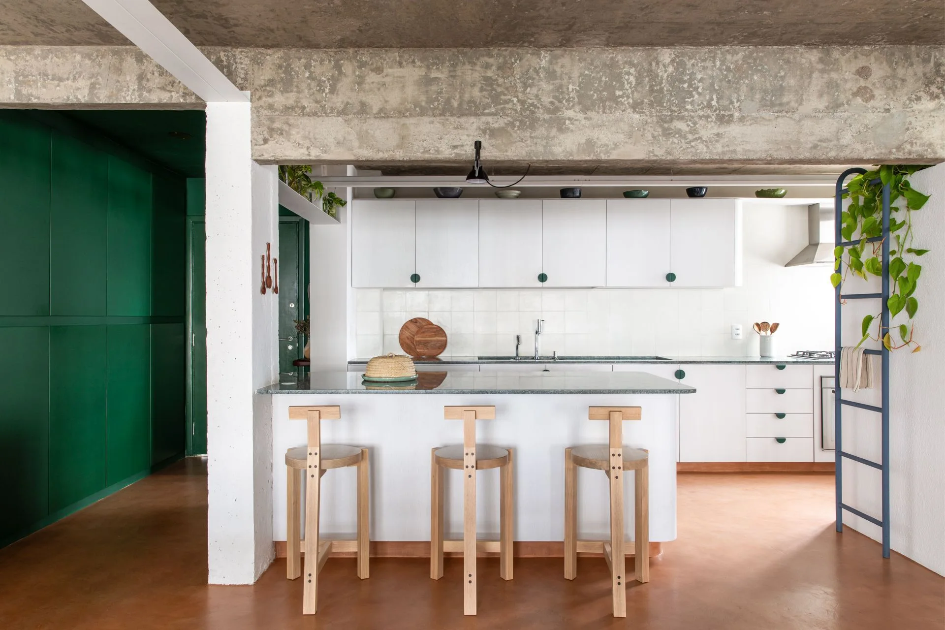 A modern kitchen with a white island, wooden bar stools, a raw concrete ceiling beam, terracotta flooring, and a deep green wall.