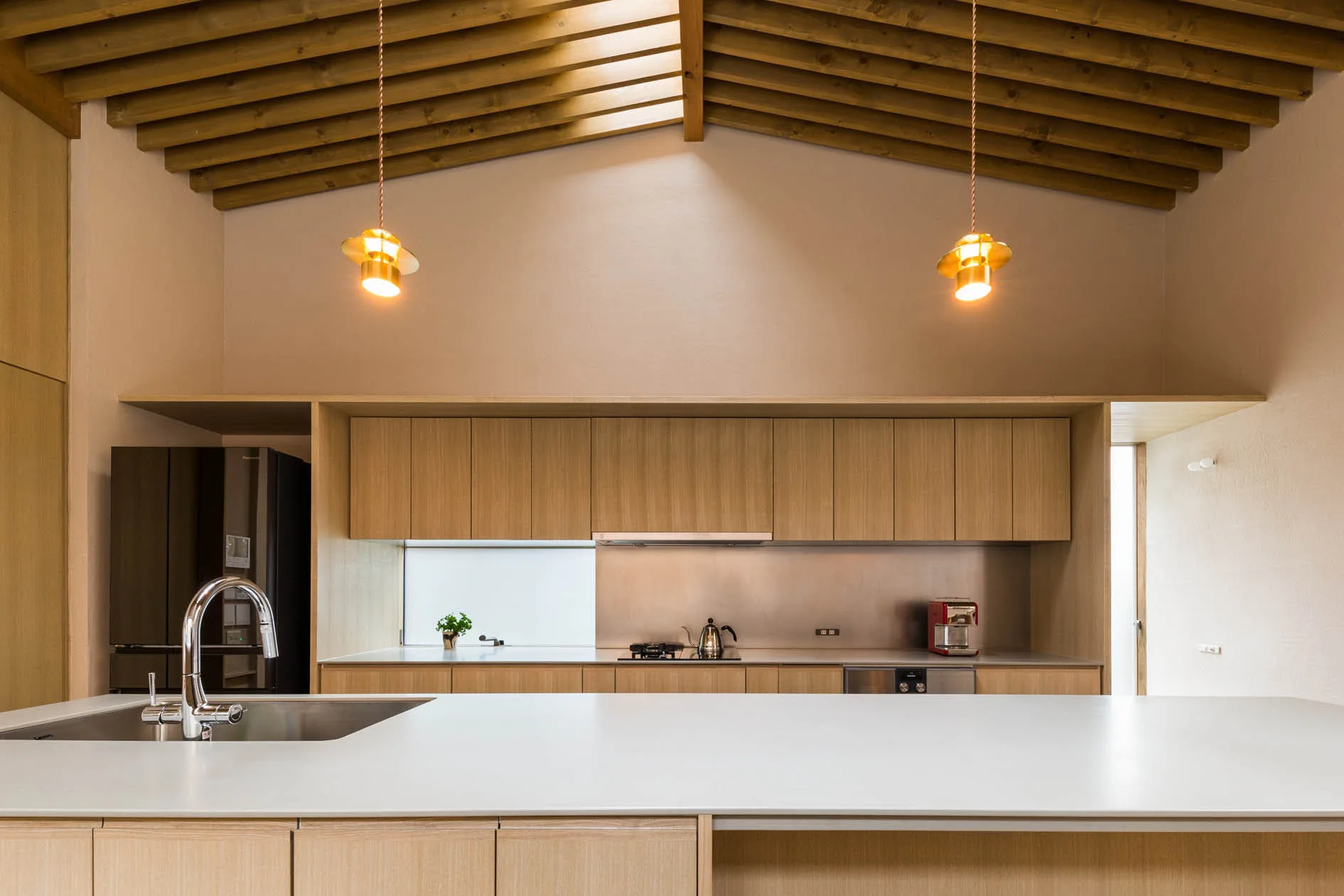 A minimalist kitchen features light wood handleless cabinetry, a white island with a sink, two gold pendant lights, and an exposed wooden beam ceiling.