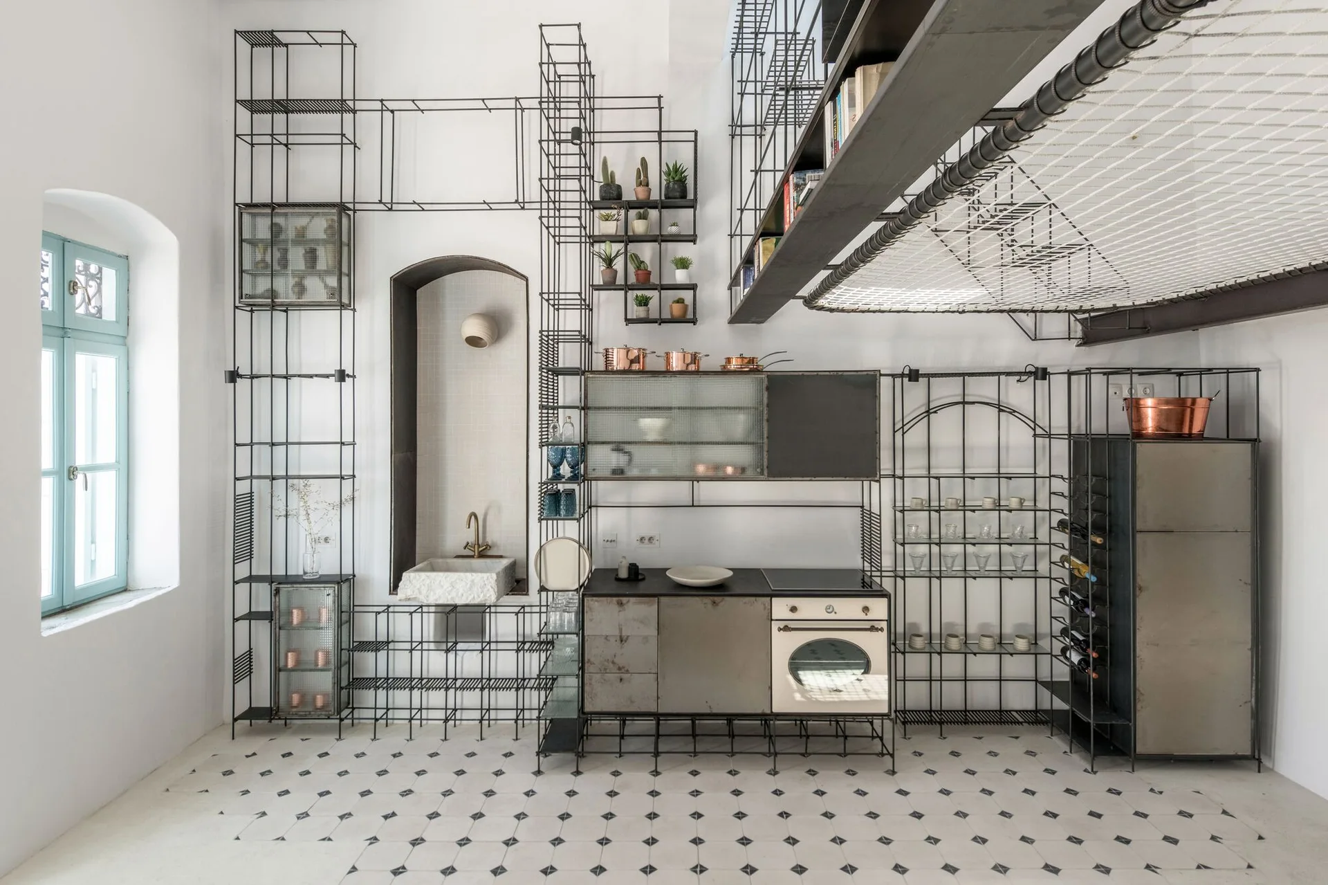 An industrial kitchen features extensive black wire-frame shelving along a white wall, a stone sink in an alcove, and a geometric tiled floor.