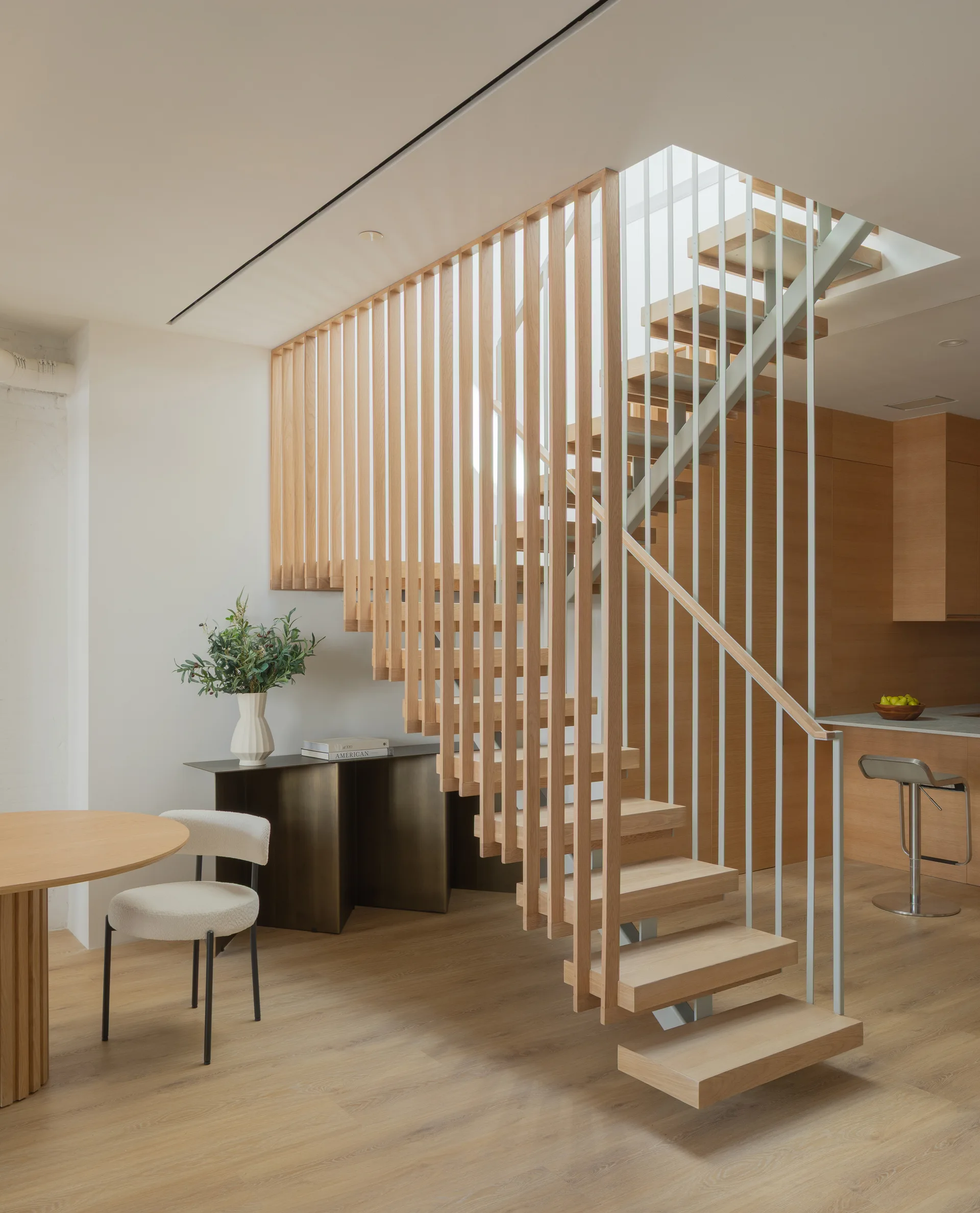 A minimalist interior features a floating wooden staircase with a vertical slat railing, a dark console table, and a white upholstered chair.