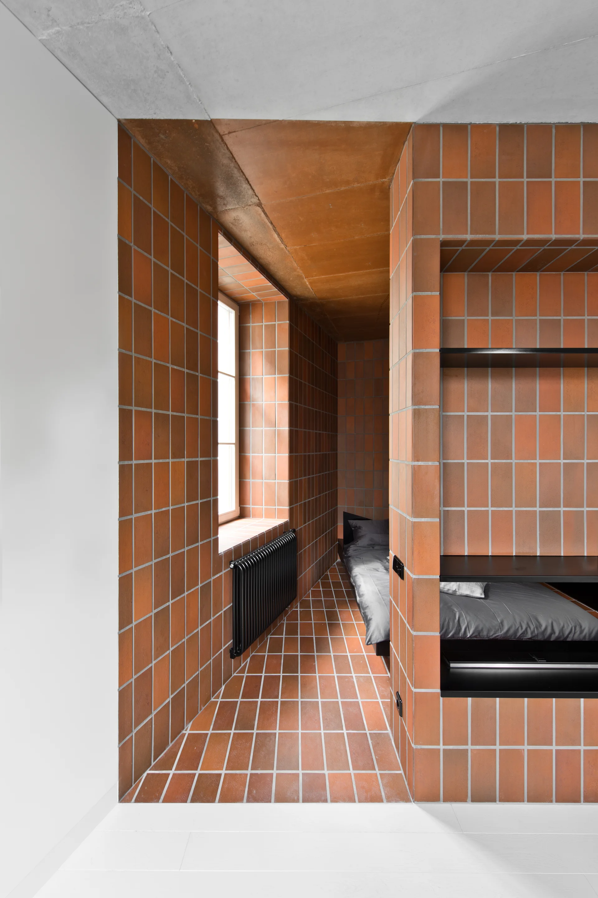 A minimalist bedroom nook wrapped in square terracotta tiles featuring a low bed, a black wall radiator, and recessed matte black metal shelves.