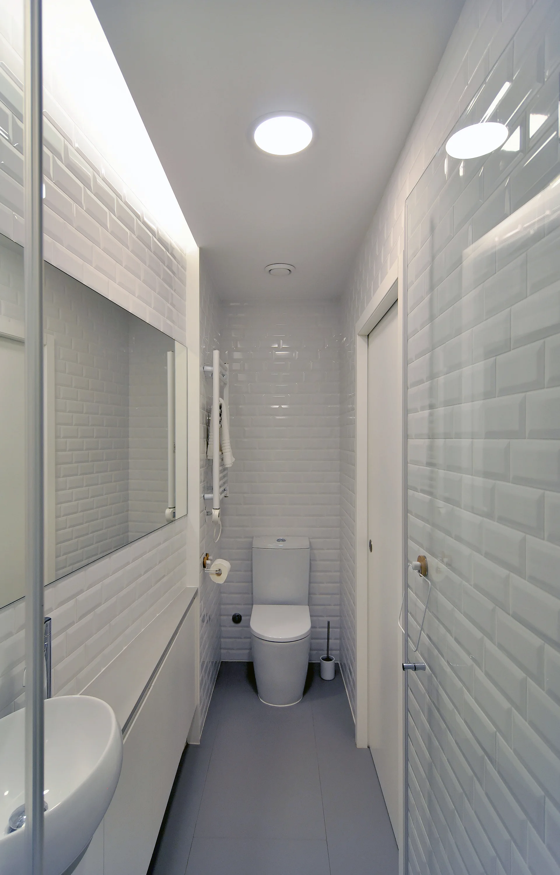 A narrow minimalist bathroom with white subway tiled walls, grey flooring, a white toilet at the far end, and a modern sink.