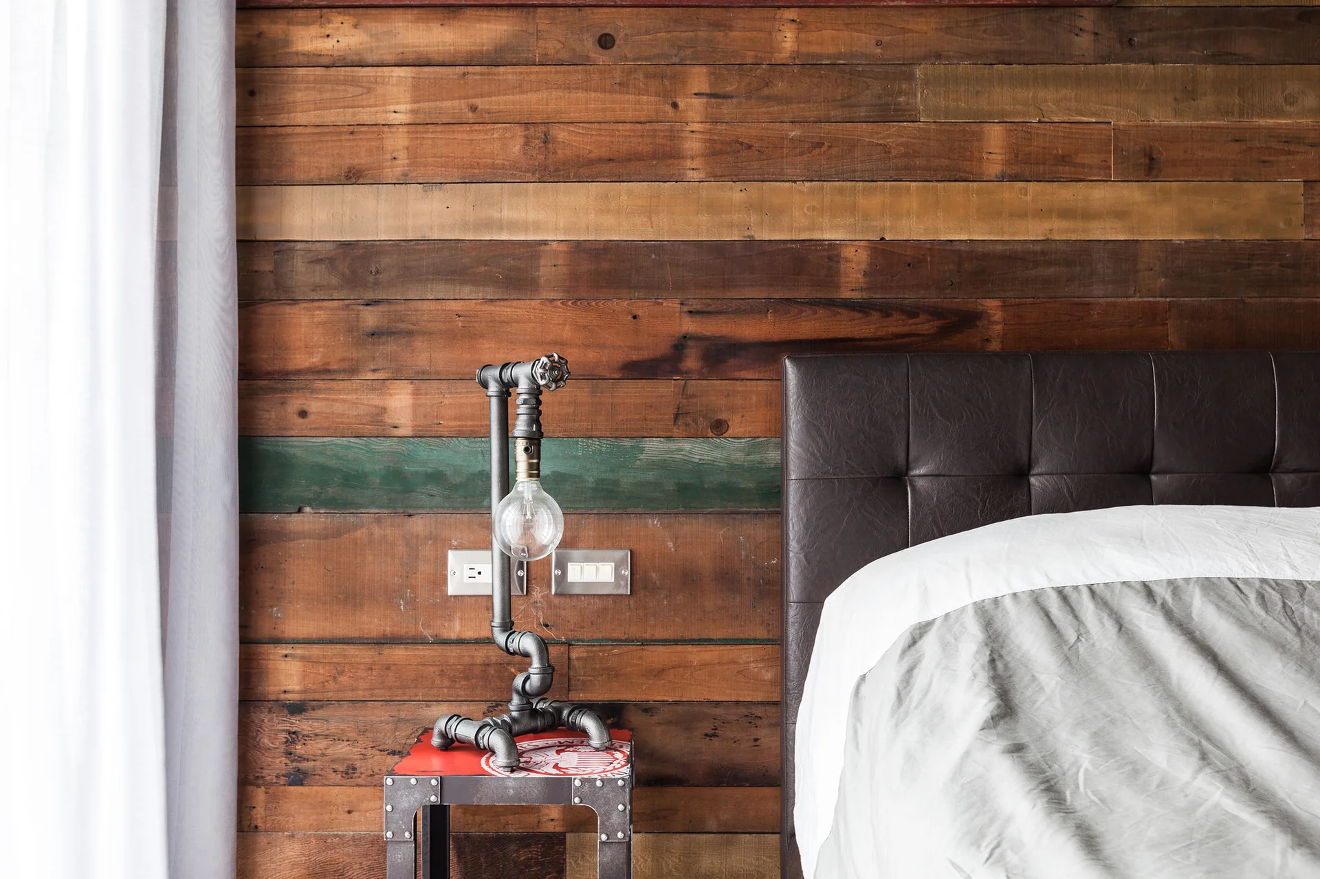 A bedroom corner features a dark tufted headboard, a rustic wood plank wall, and an industrial pipe lamp on a small bedside table.