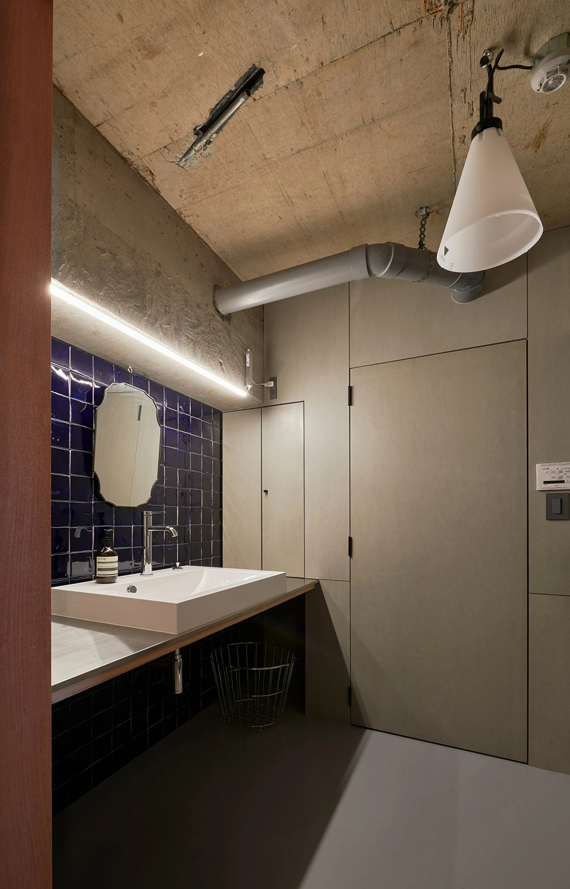 A modern bathroom features a white sink against glossy navy blue tiles, flat grey wall panels, and an unfinished concrete ceiling.
