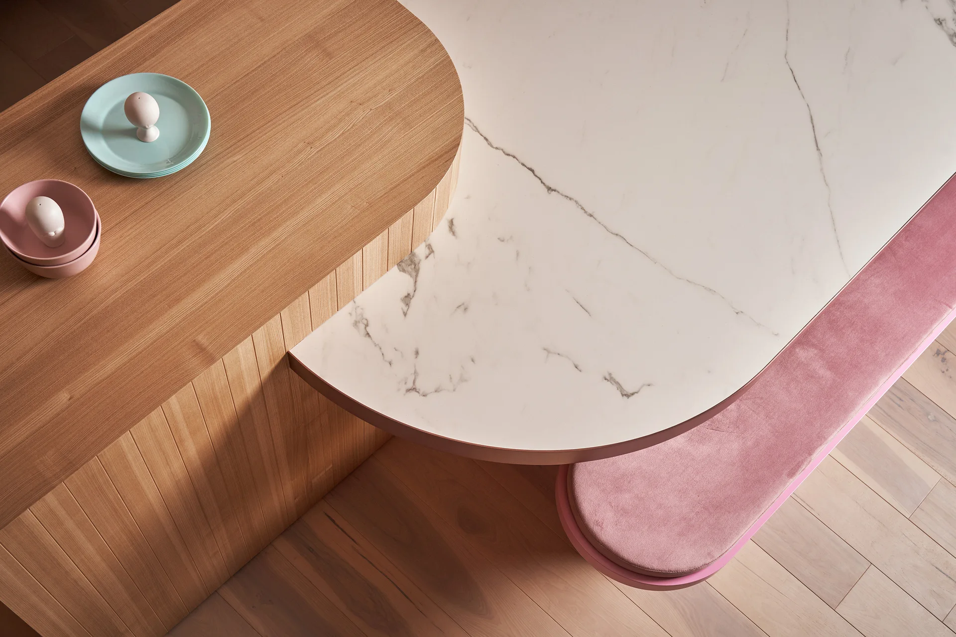 A top-down view of a curved white marble dining table connected to a fluted wood island and a pink velvet upholstered bench.