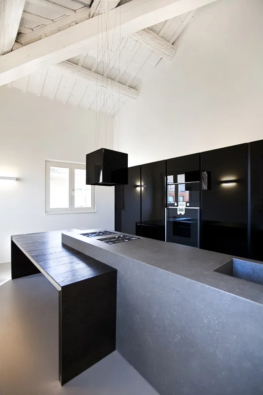 A minimalist kitchen featuring a grey stone island with an attached dark wood table, glossy black cabinets, and white painted ceiling beams.