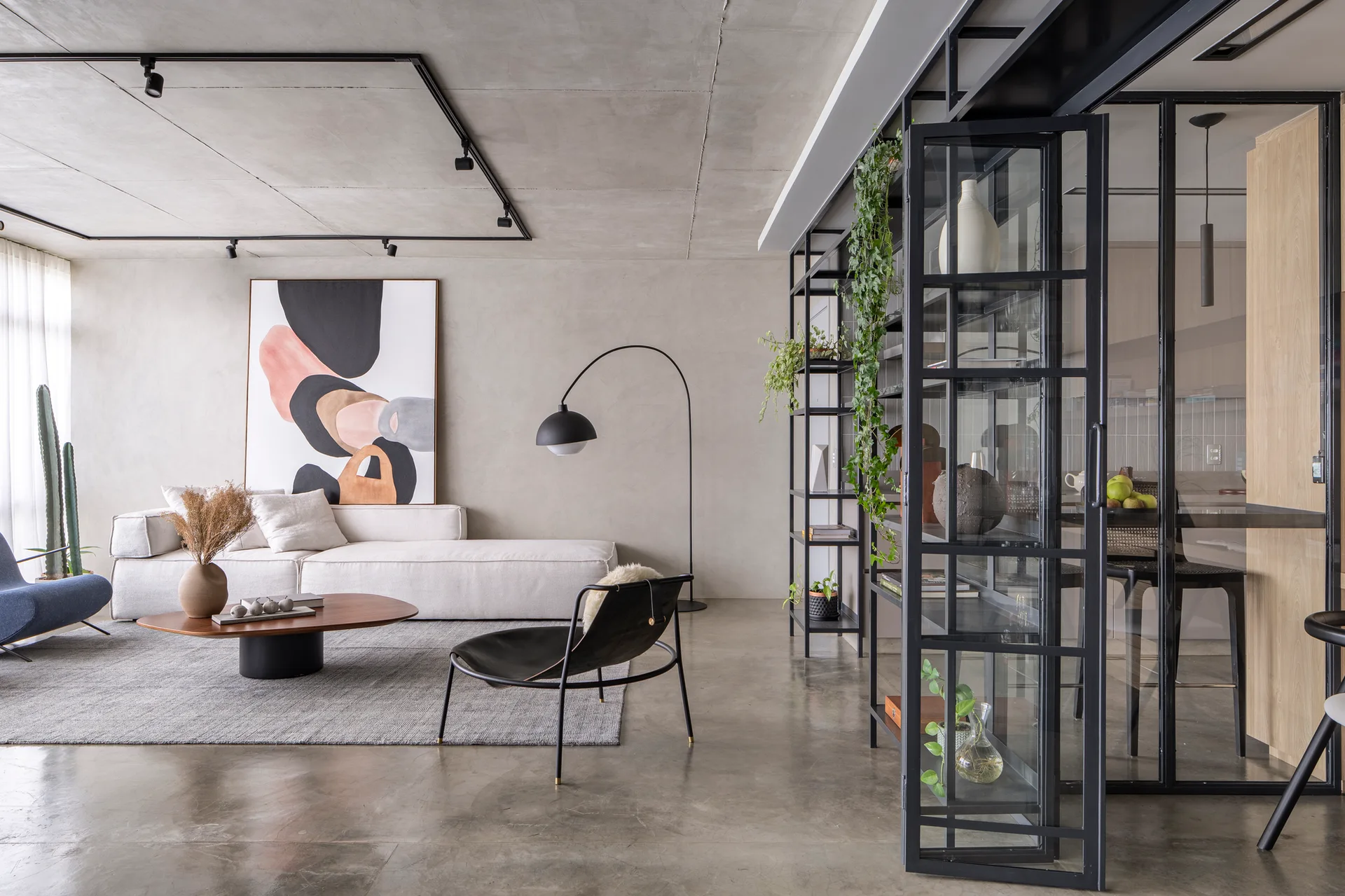 A modern industrial living room features a low white sofa, concrete floors and ceiling, and a black metal-framed glass partition.