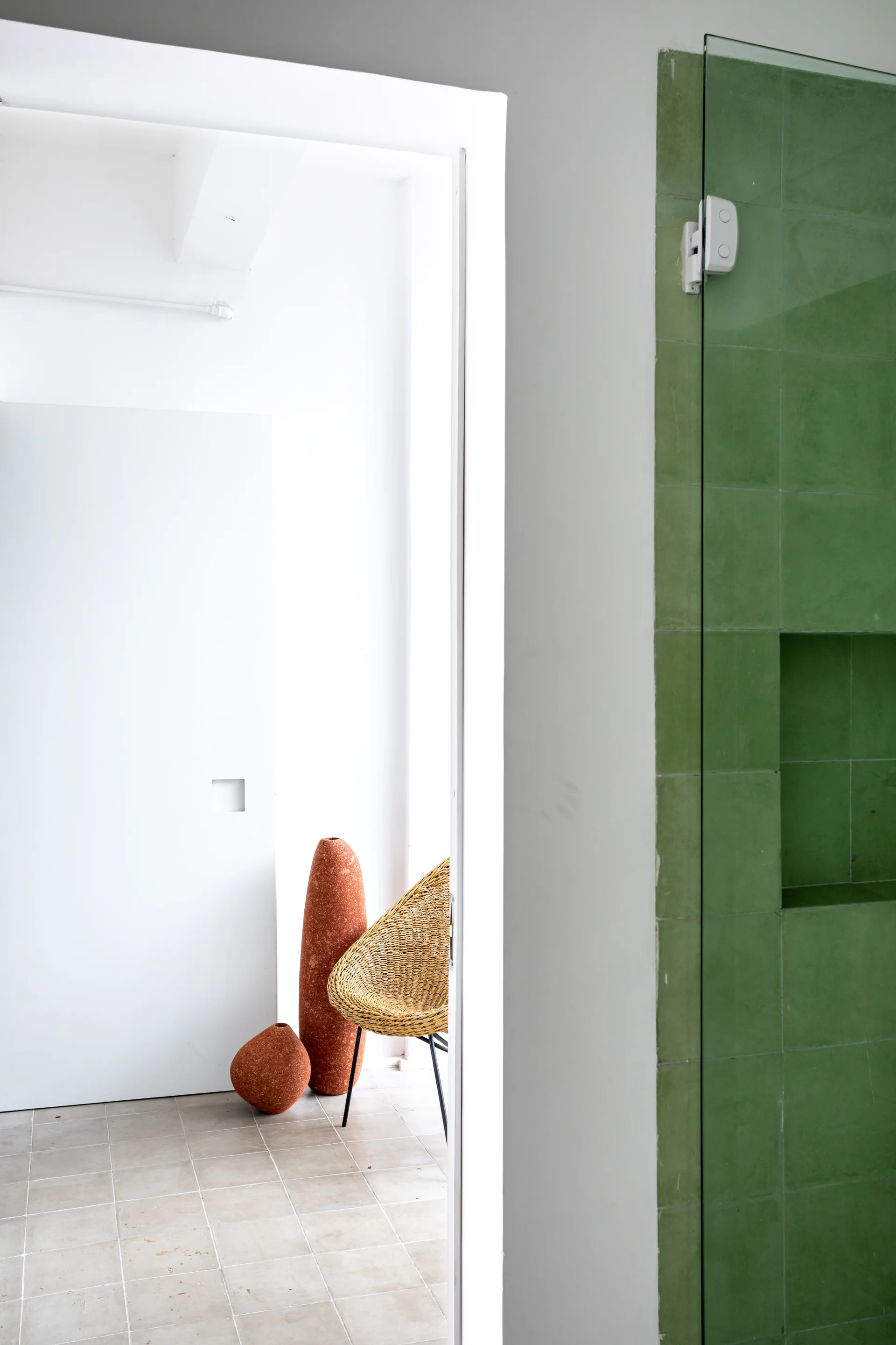 A view through a white doorway shows a green tiled shower wall next to an adjacent room with a wicker chair and two terracotta vases.