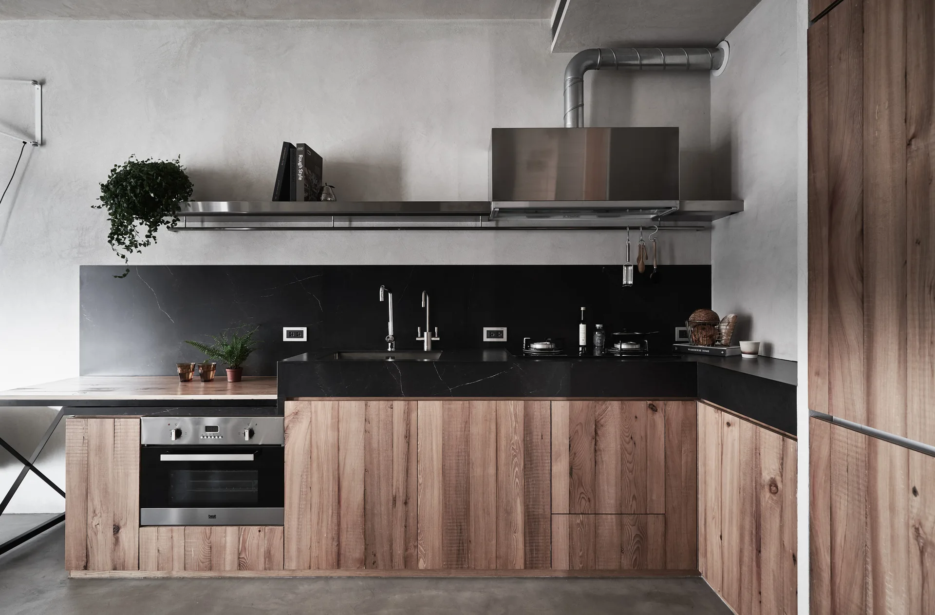 A modern industrial kitchen with natural wood lower cabinets, a black stone countertop and backsplash, a stainless steel hood, and grey plaster walls.