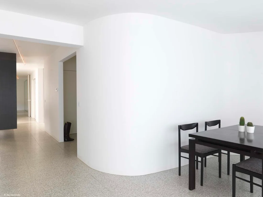 A dark wood dining table and chairs stand next to a large white curved partition wall on speckled grey flooring leading to a hallway.
