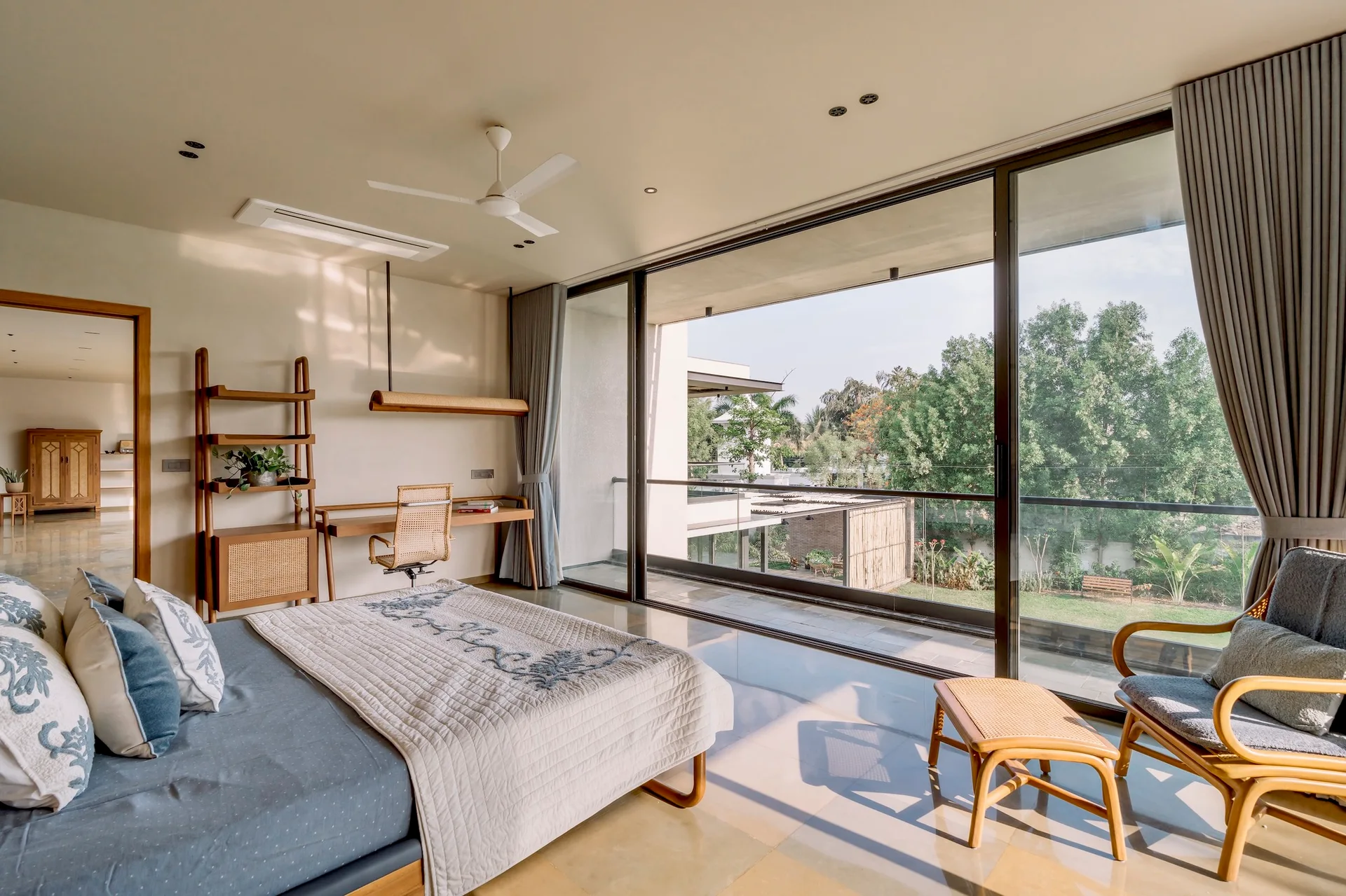 A sunlit bedroom features a bed with blue bedding, a wooden writing desk, cane chairs, and large sliding glass doors opening to a lush balcony.