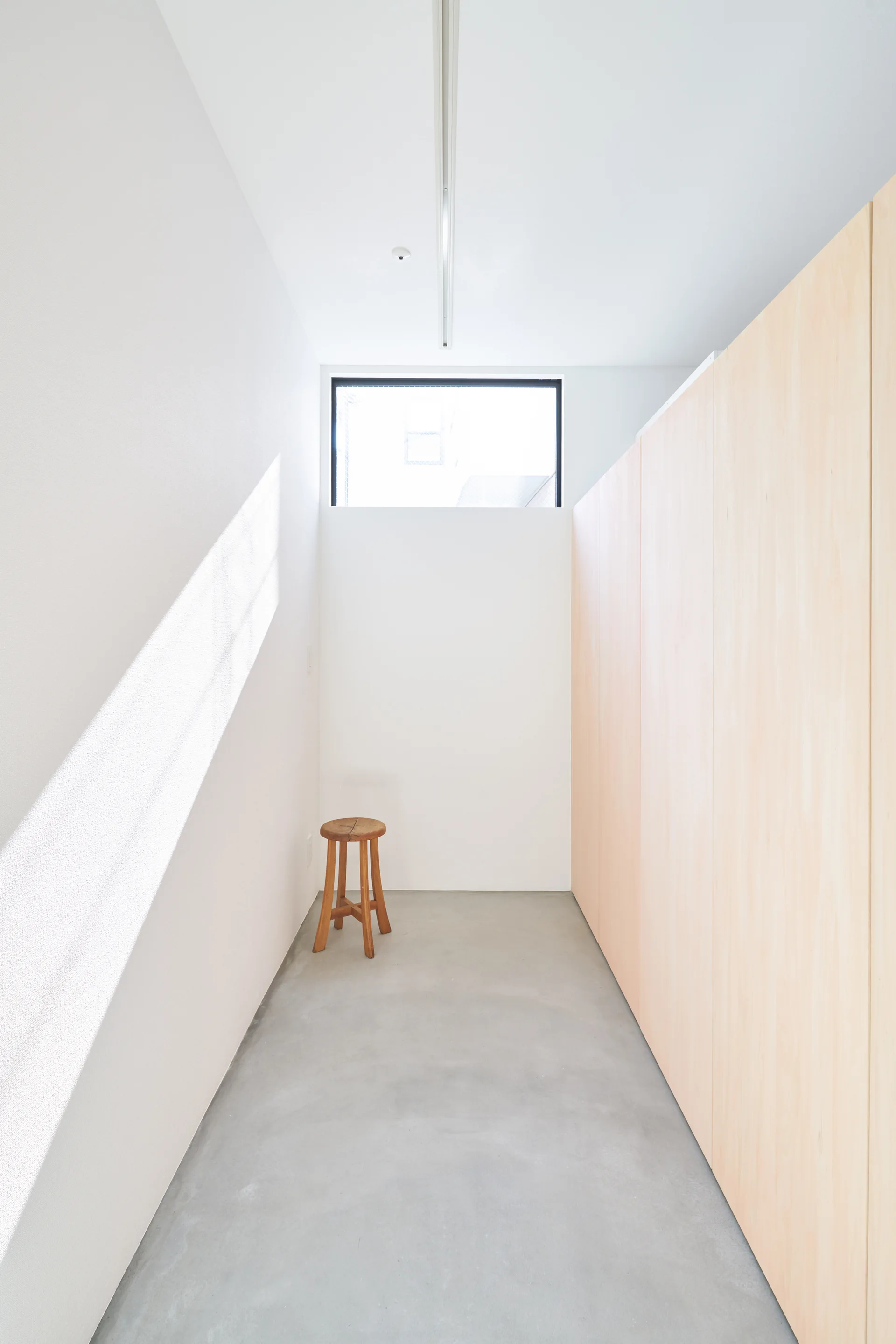 A minimalist hallway with concrete floors, white walls, full-height light wood wall panels, and a single wooden stool under a high rectangular window.