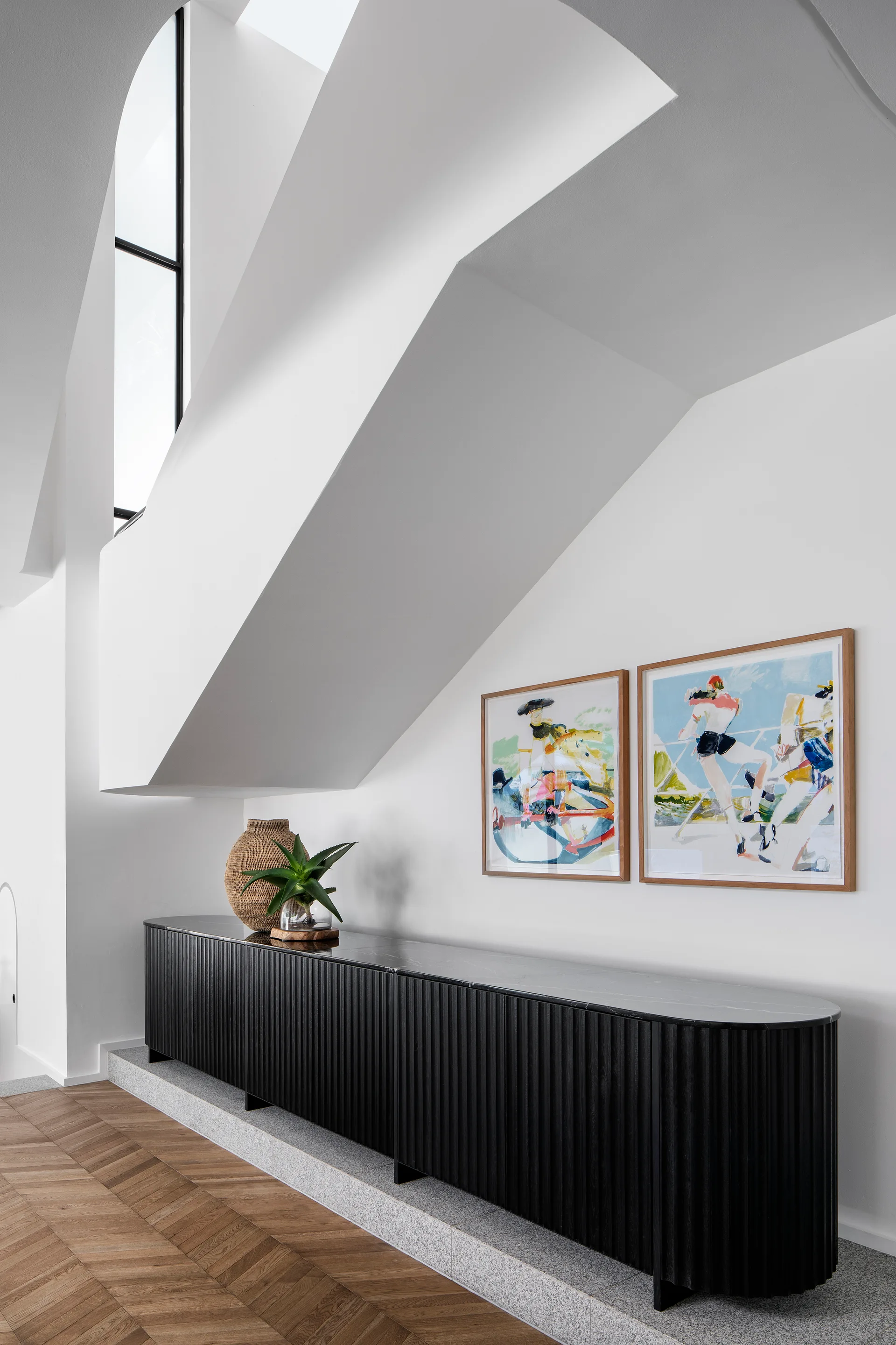 A modern white hallway features a black fluted sideboard console beneath sculptural ceiling angles and framed art prints.