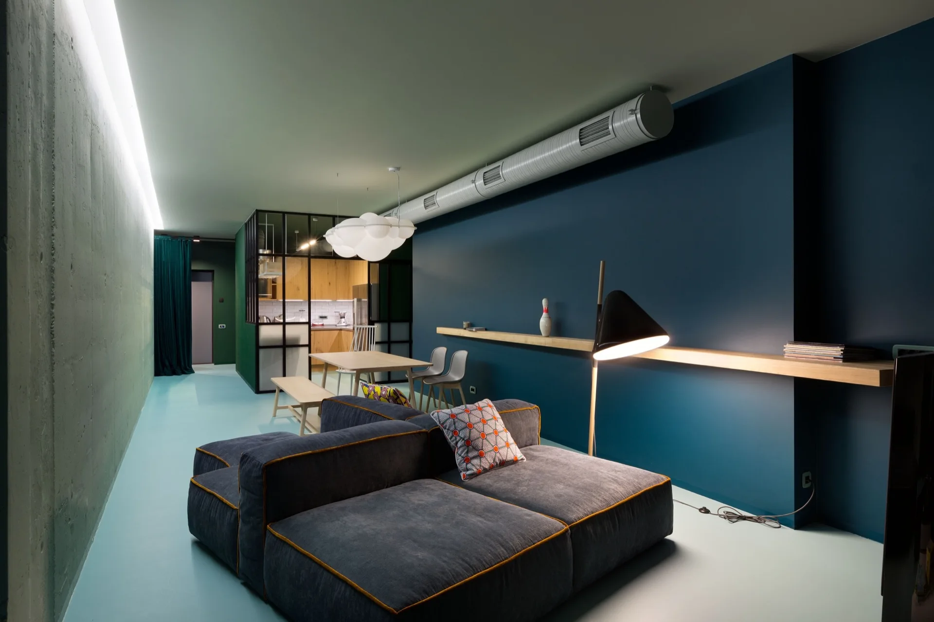 A low-profile modular sofa sits on a light blue floor in a modern room with a dark teal wall, a long floating wood shelf, and exposed ceiling ducts.