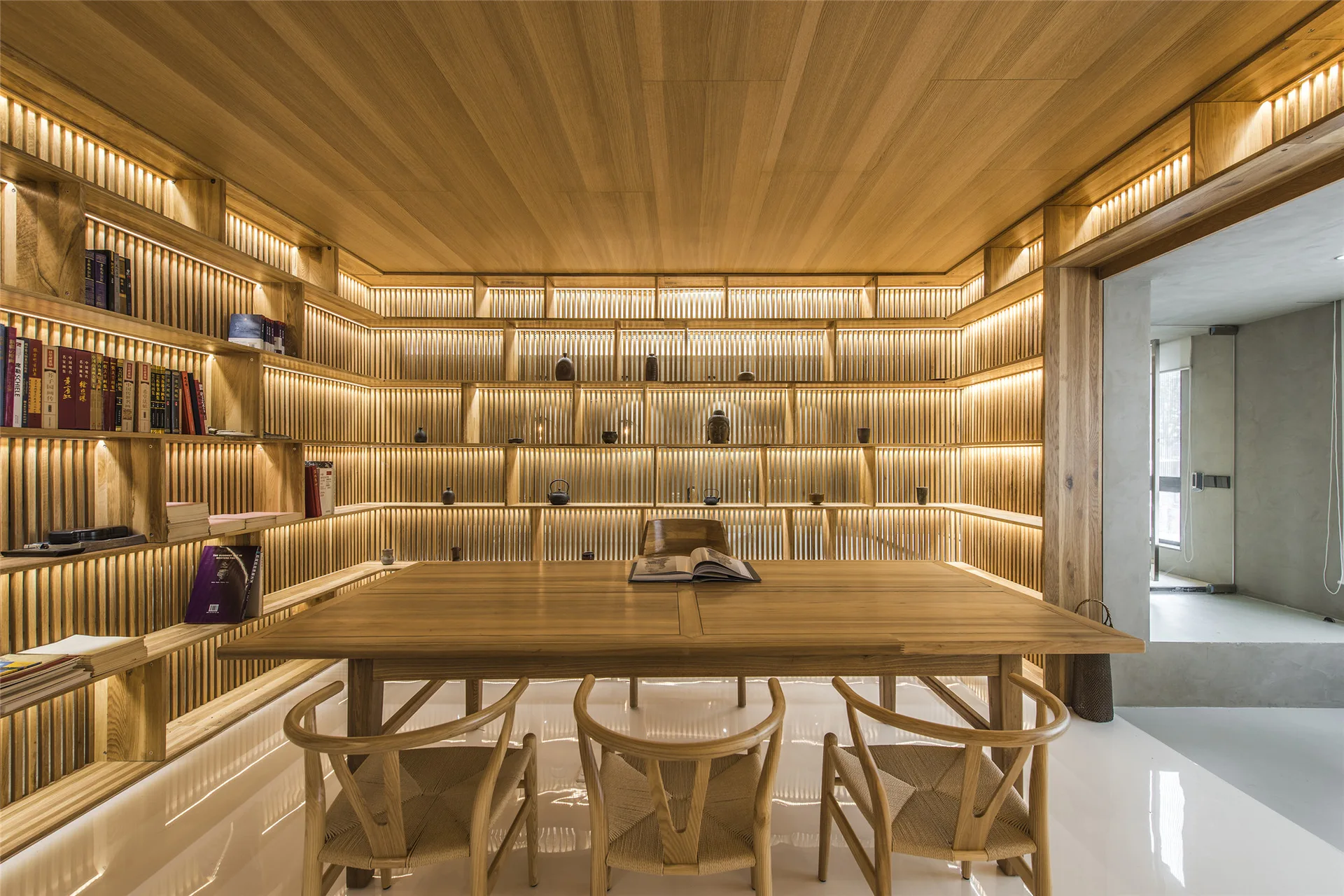 A minimalist library features a wood table with chairs and wrap-around wood shelving with integrated warm lighting on slatted walls.