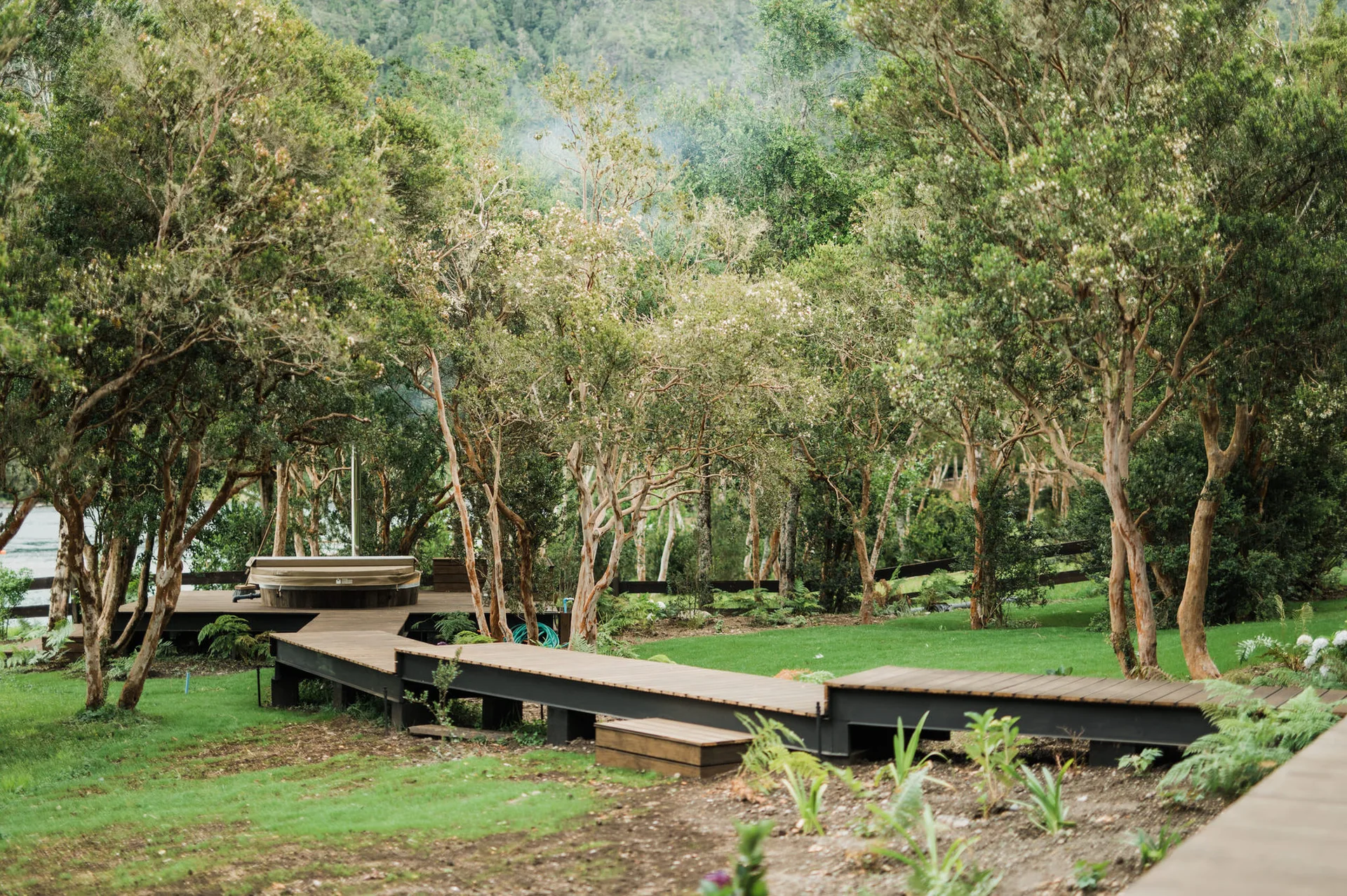 A raised wooden boardwalk winds through a grassy lawn with tall trees, leading to a circular hot tub on a platform deck.
